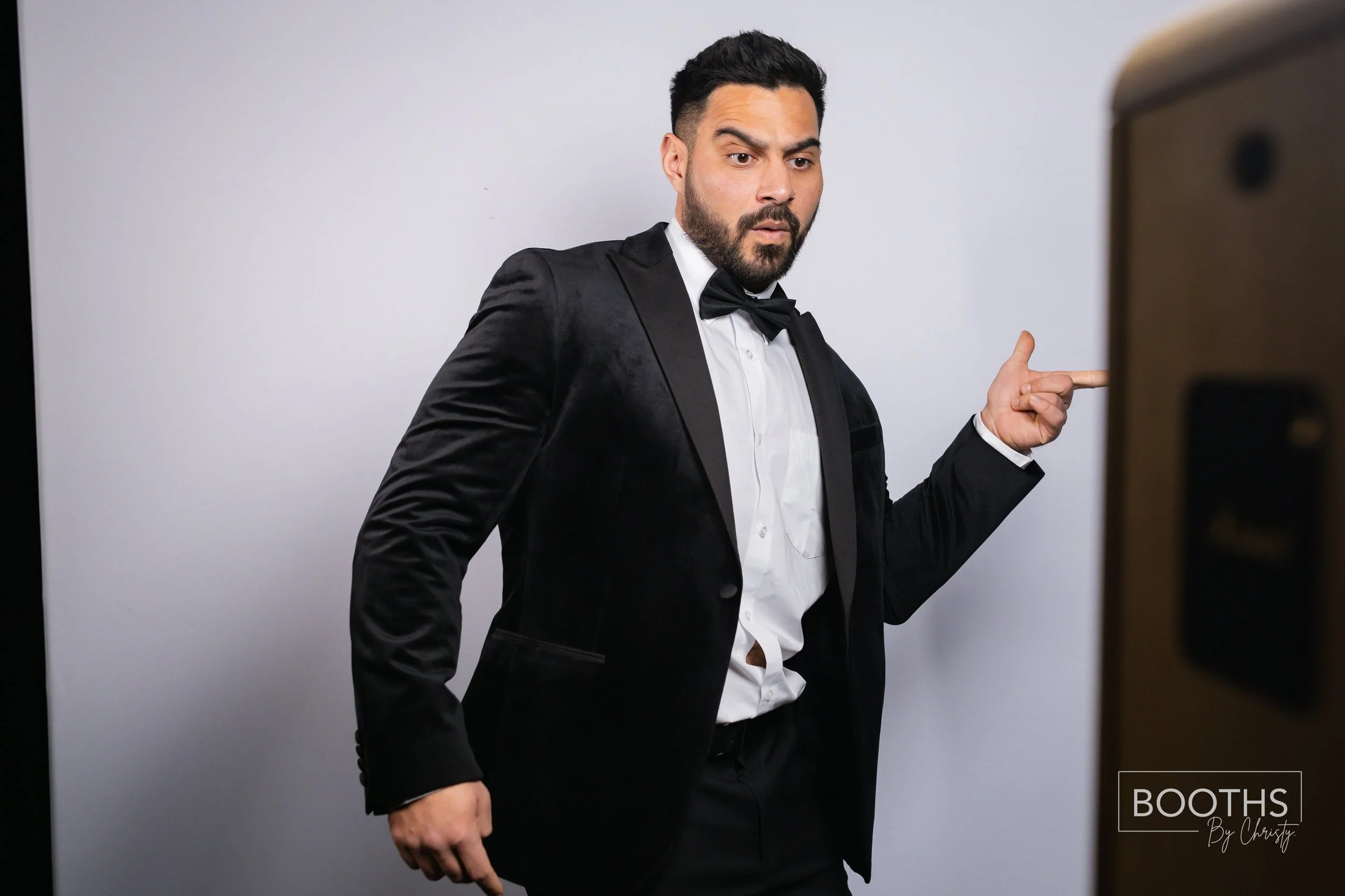 Man in black tuxedo with white shirt and black bow tie pointing at a mirror or screen against a plain background.