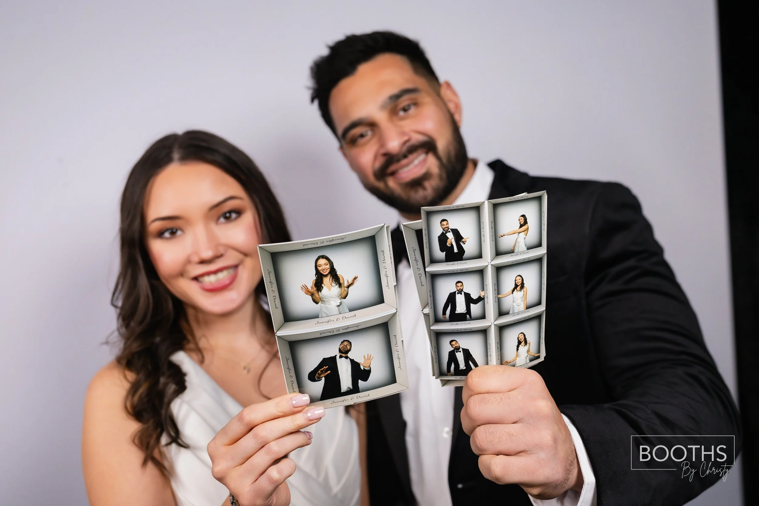 A smiling woman and man at a wedding or event holding a photo booth strip with multiple pictures of themselves posing with playful gestures. The woman has long dark hair and wears a sleeveless white dress, while the man has short dark hair, a beard,