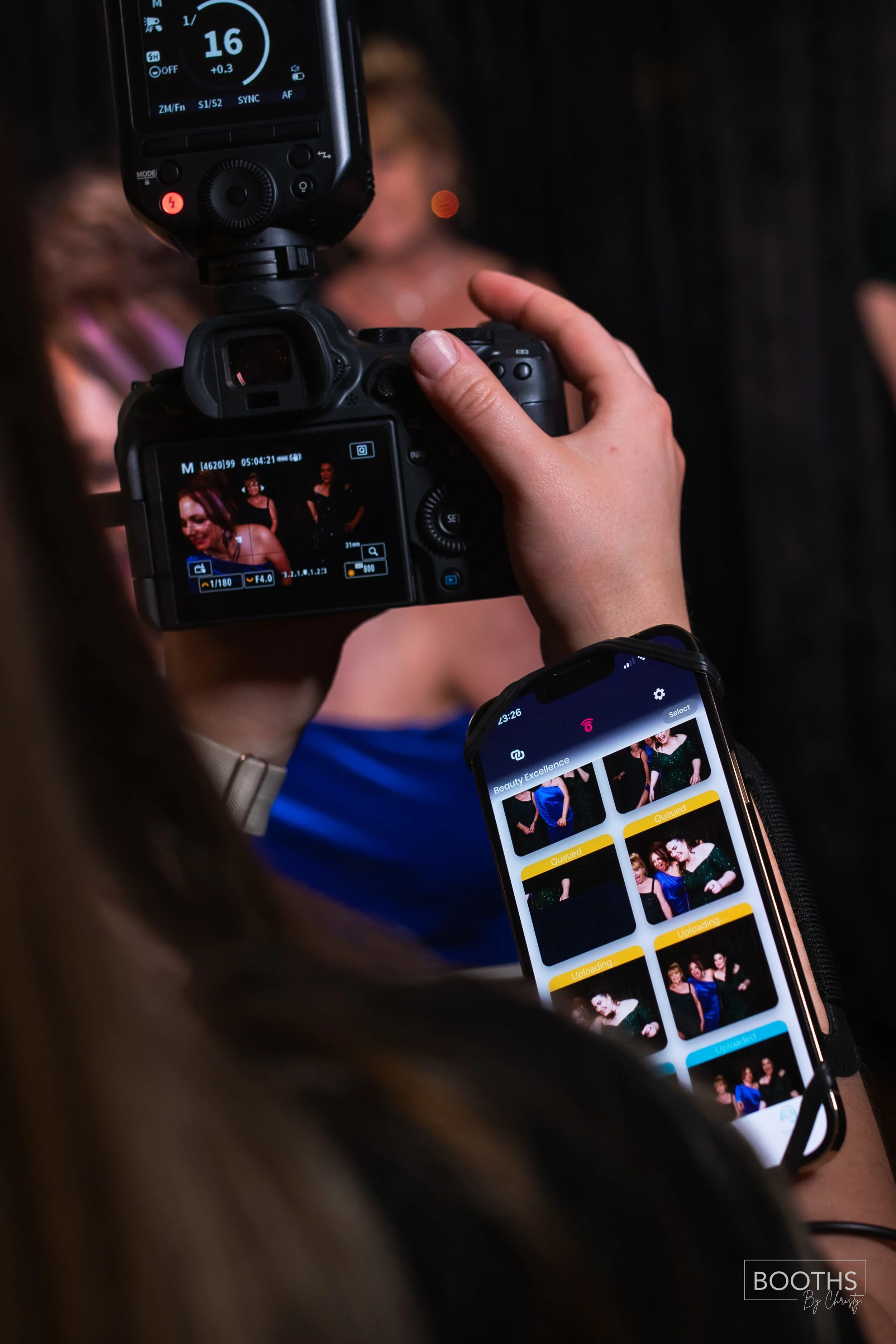 Photographer taking pictures of women at a photo booth event with a DSLR camera and a smartphone. The camera display shows the women posing, with a background of black curtains. The smartphone screen shows a gallery of candid photos of the women.
