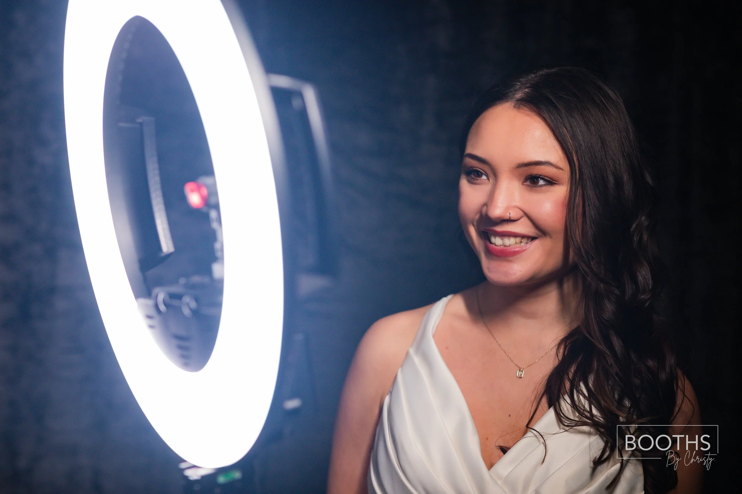 A young woman with dark wavy hair smiling at a ring light in a photo booth.