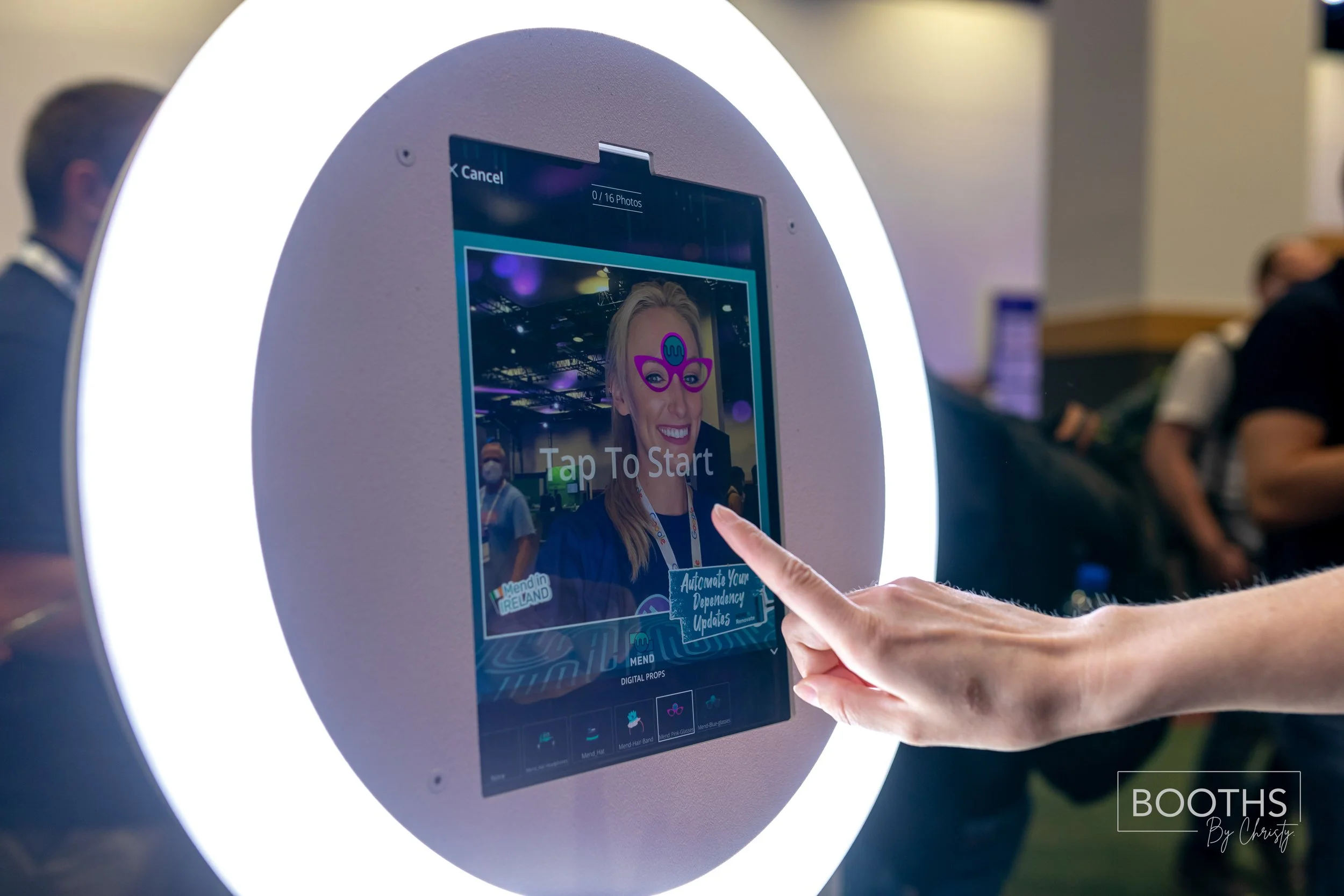 A person is using a digital photo booth with a touchscreen interface surrounded by a circular light at an event. The screen displays a woman smiling with digital glasses and text that says "Tap To Start."