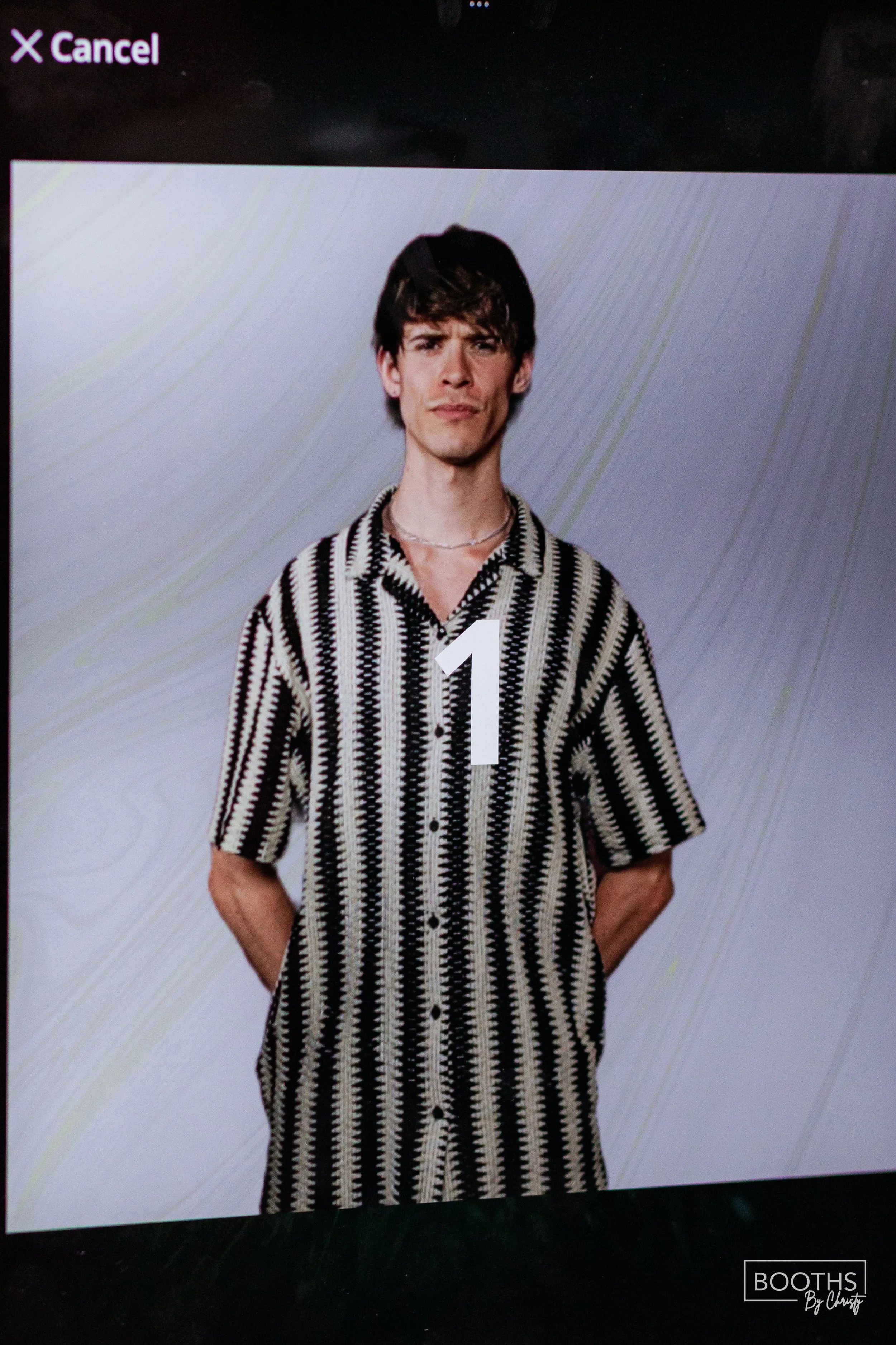 A young man with dark, slightly tousled hair and a serious expression, wearing a black and white patterned short-sleeve button-up shirt, with a large white number 1 overlaid on him, standing against a plain background.