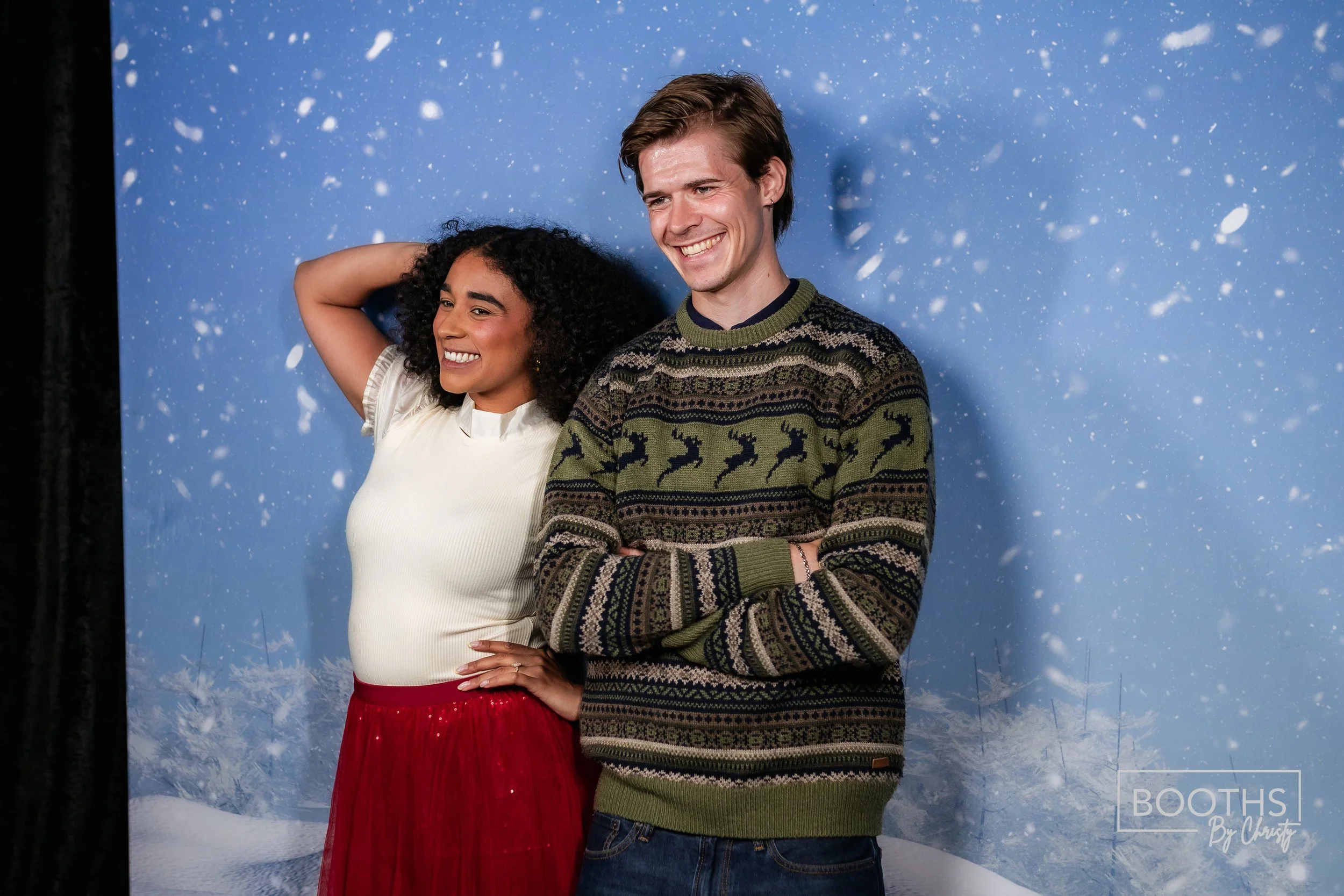 Two people, a woman and a man, standing in front of a winter-themed backdrop with snow, smiling. The woman wears a white top and a red skirt, and the man wears a multicolored sweater with a reindeer pattern.