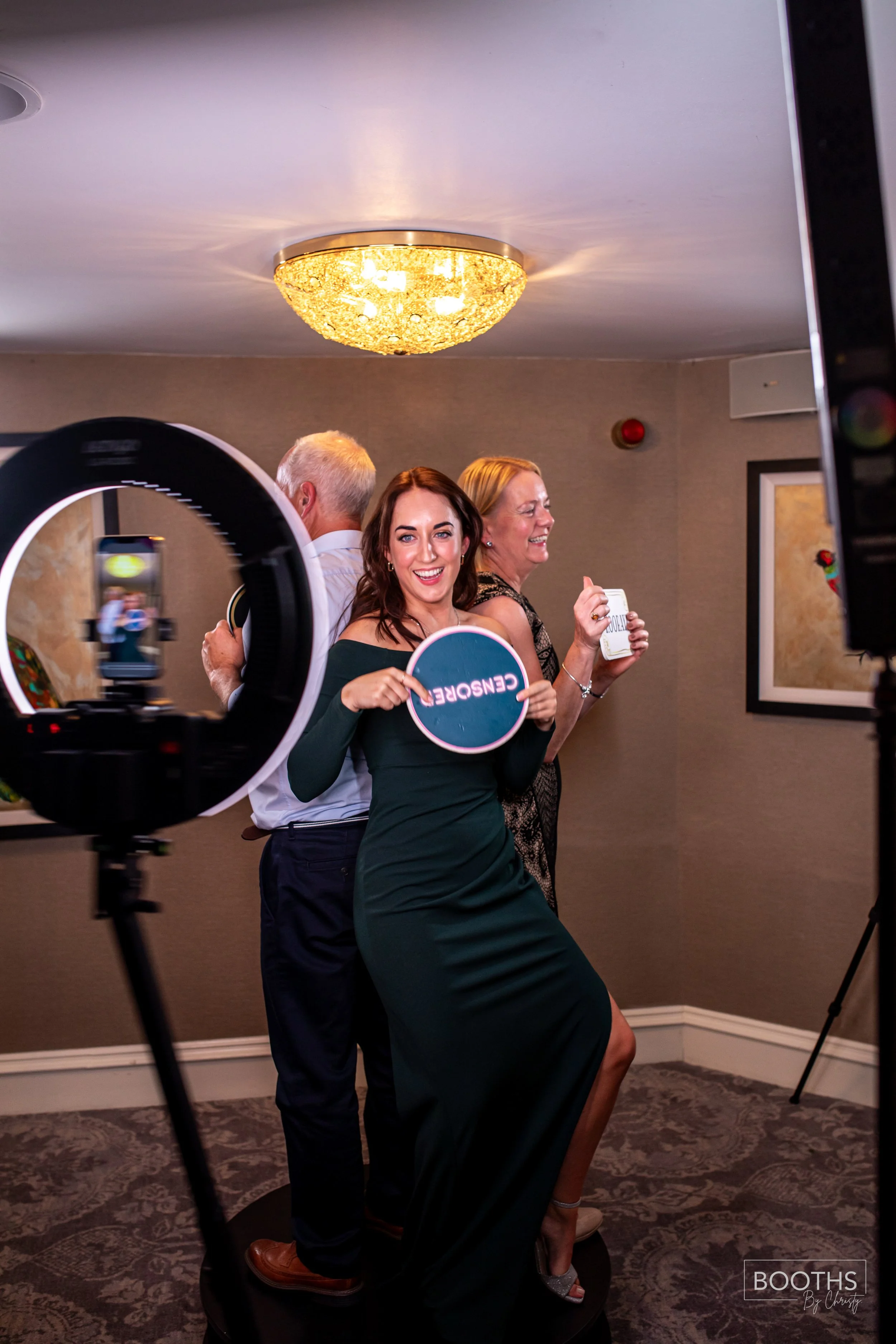 Three people, two women and a man, at a photo booth, with the woman in the front holding a sign that says 'CENSORED,' smiling and posing. The woman on the right is holding a mug and laughing, while the man behind has his face turned away. The photo i