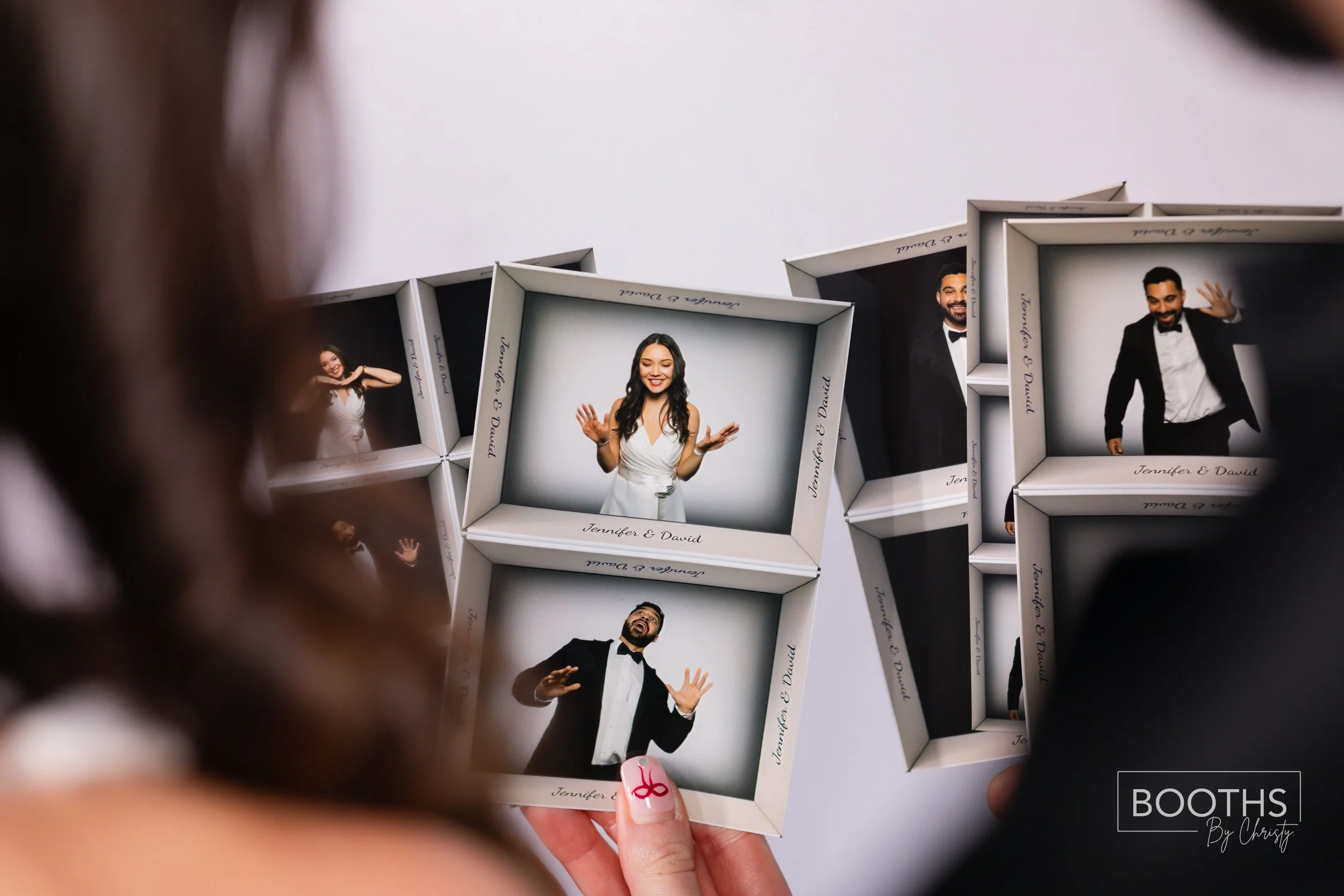 Close-up of a hand holding a photo booth picture of a woman in a white dress and a man in a tuxedo, with other similar pictures in the background.