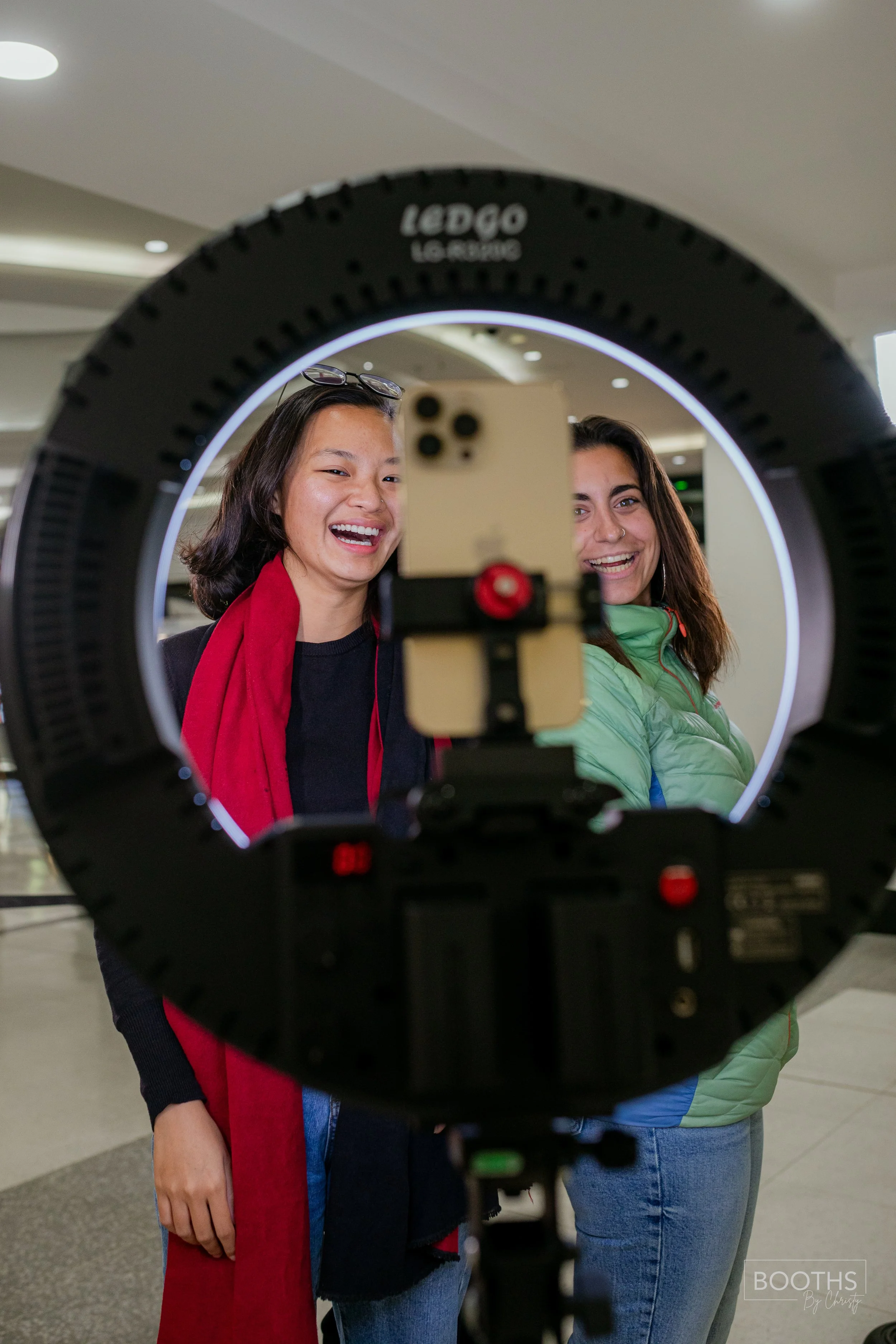 Two women taking a photo together in a shopping mall, seen through a ring light and smartphone setup.
