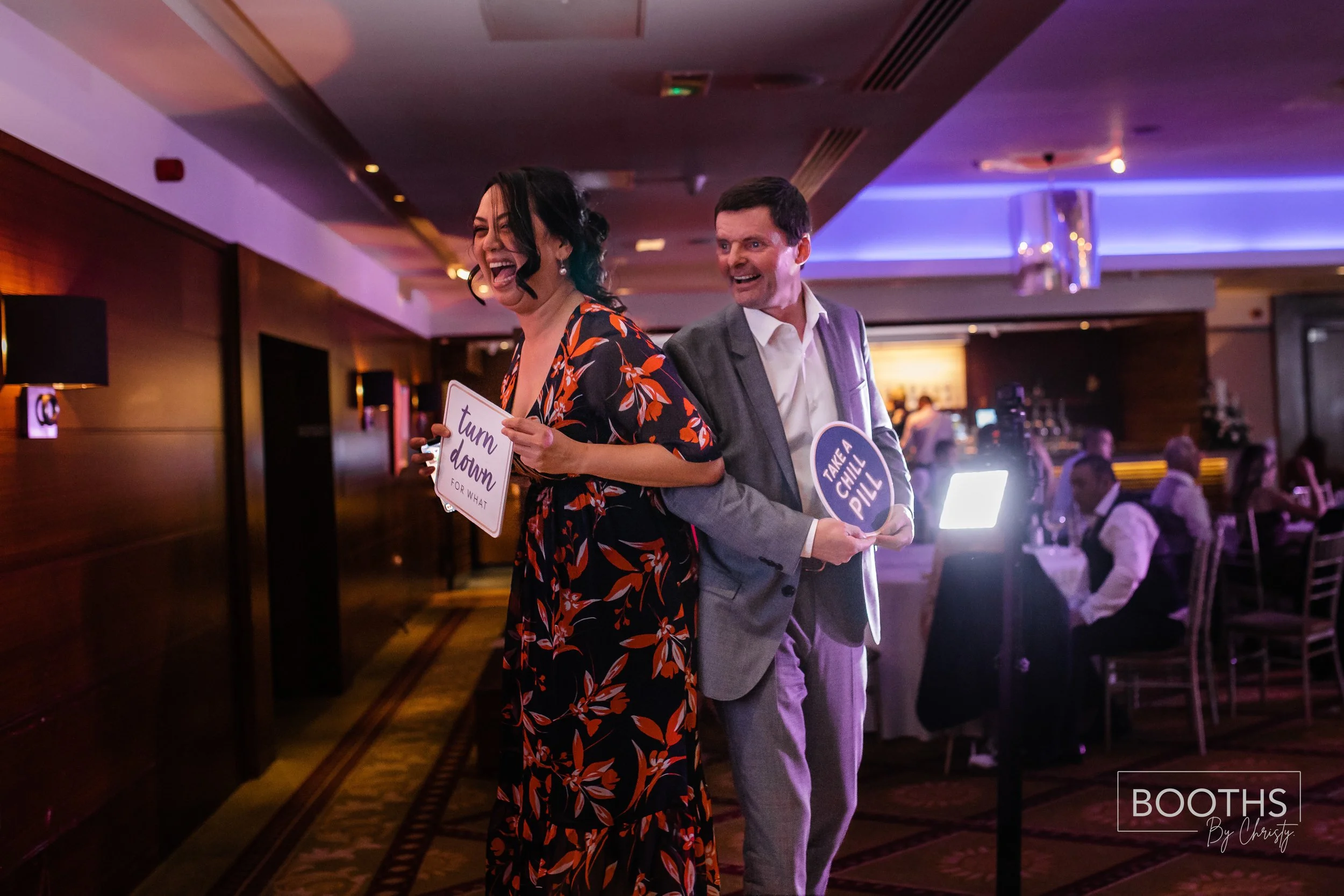A woman and a man at an indoor event, standing back to back, holding signs that read "turn down for what" and "take a chill pill" respectively. The woman is laughing and wearing a floral dress, while the man is smiling and dressed in a suit. There ar