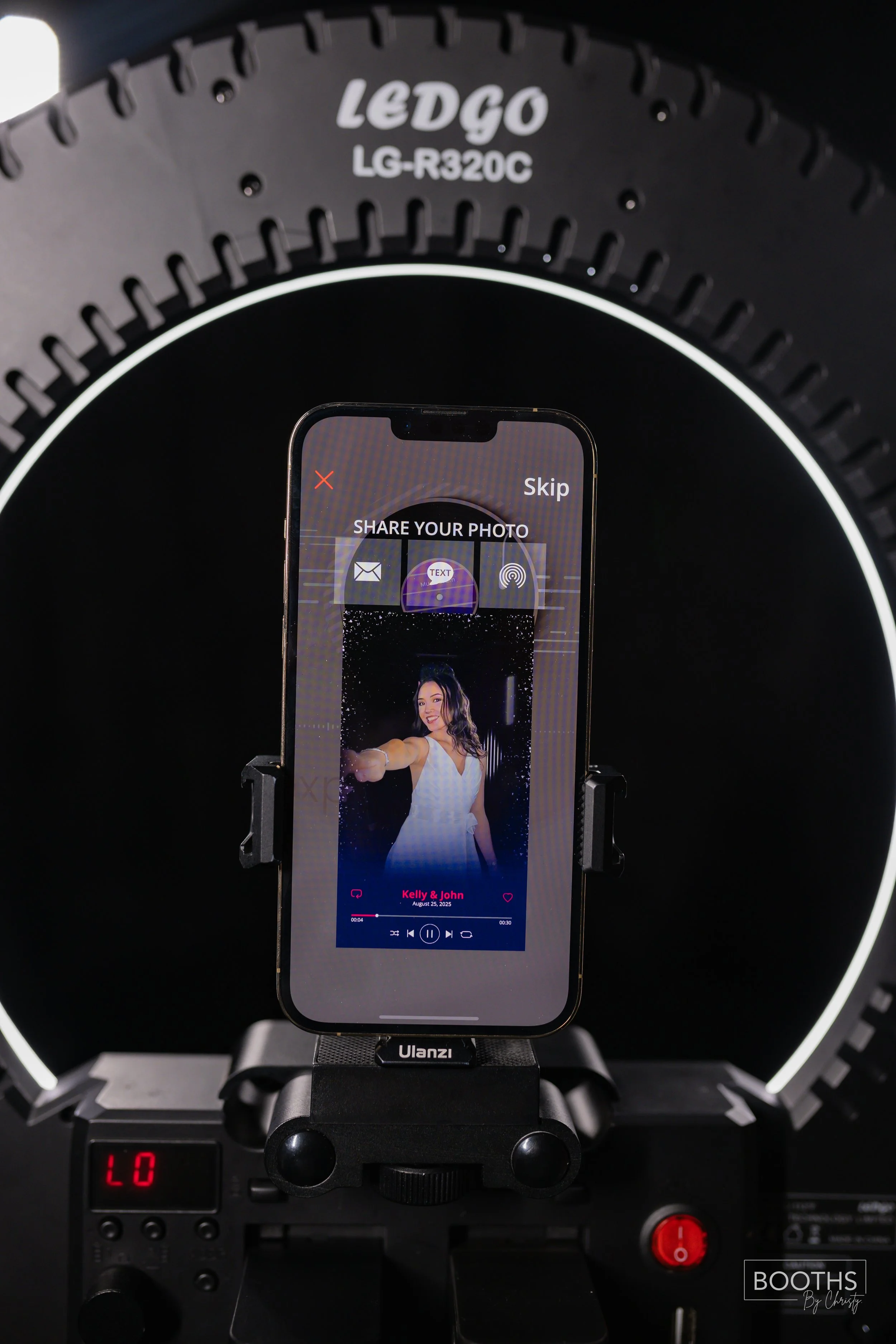 Smartphone mounted on a stabilizer shows a photo of a woman in a white dress smiling and taking a selfie, with a music player interface below her picture. The phone is positioned in front of a circular studio light and a black background.