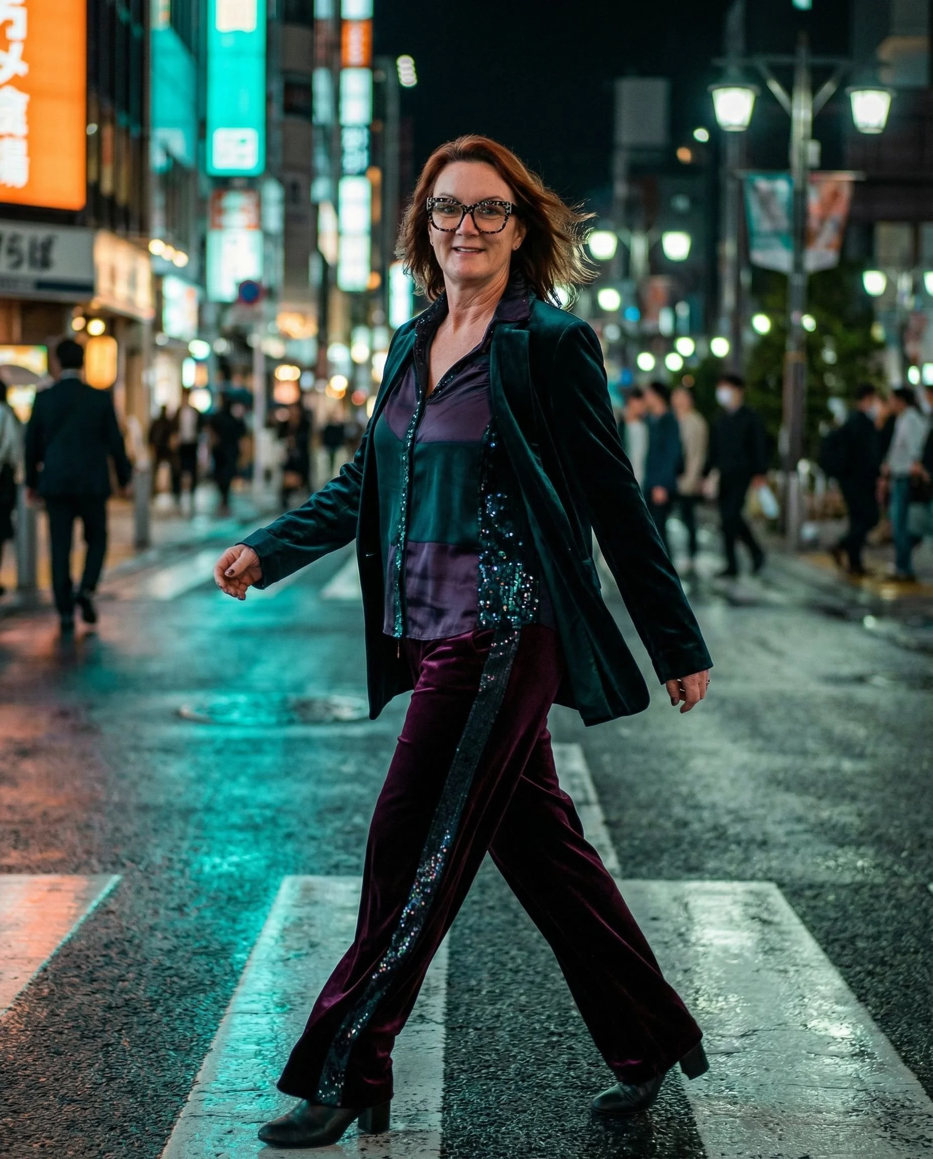 A woman crossing a busy street at night in an urban area with bright neon lights and signs, wearing a dark velvet jacket and patterned pants.