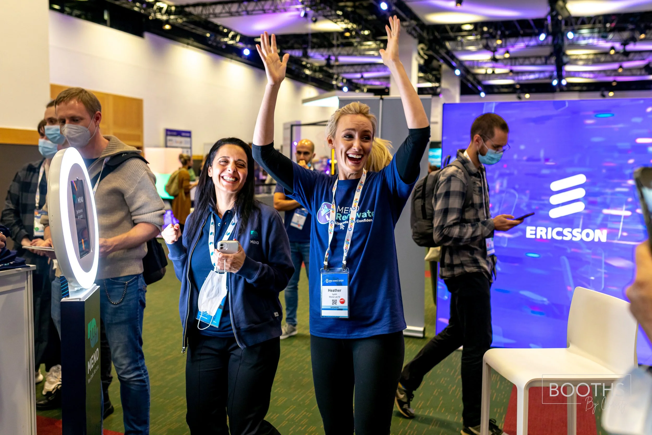 Two women, one with her arms raised in celebration and both smiling, attending a conference or trade show booth with a large screen displaying the Ericsson logo in the background. Several other attendees are seen around, some looking at their phones.
