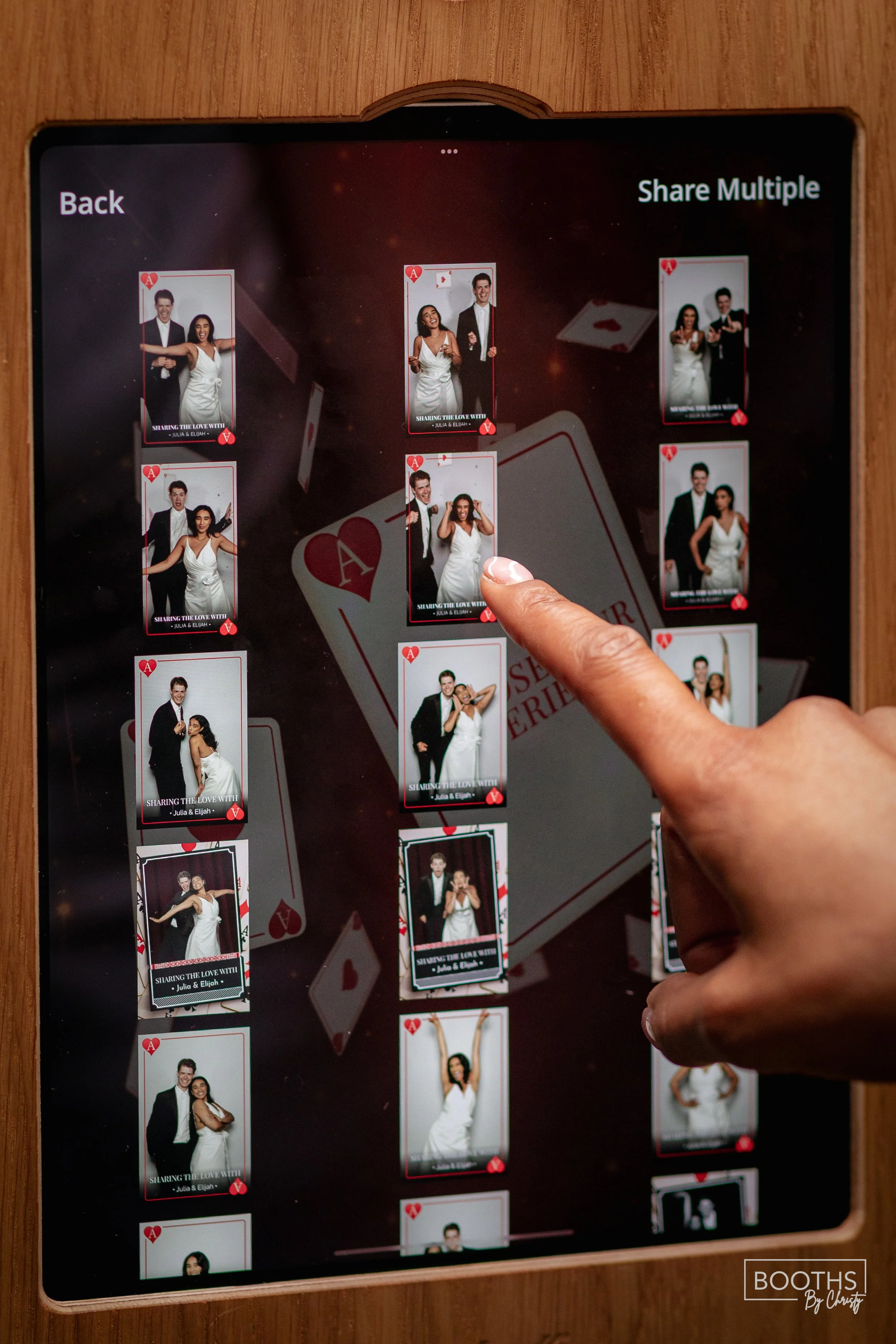 A person is selecting a photo displayed on a digital photo booth screen, which shows multiple pictures of a couple in wedding attire, with the backdrop of a deck of cards.