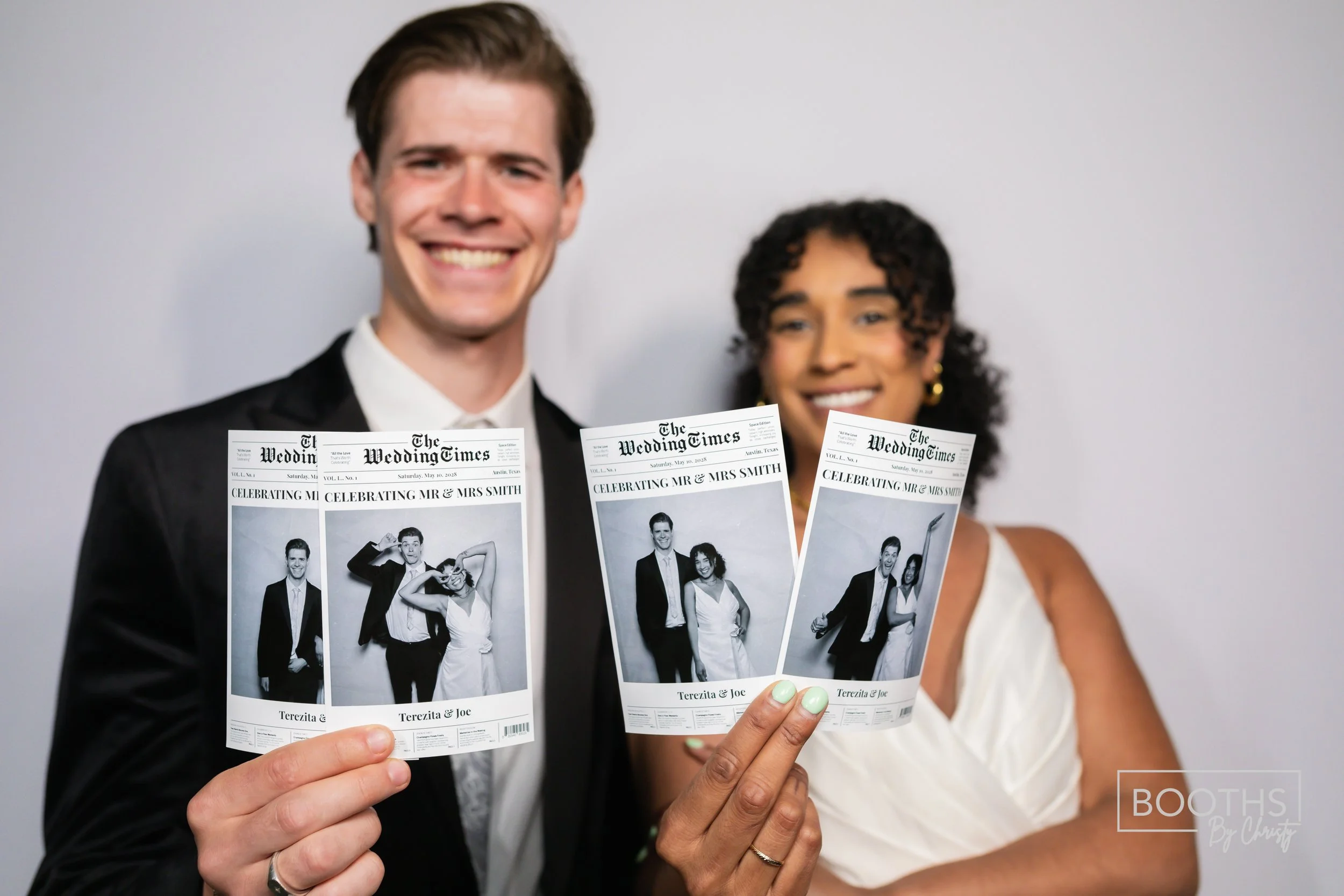 A smiling couple in wedding attire holding wedding photo booth strips with black-and-white pictures, standing against a plain background.