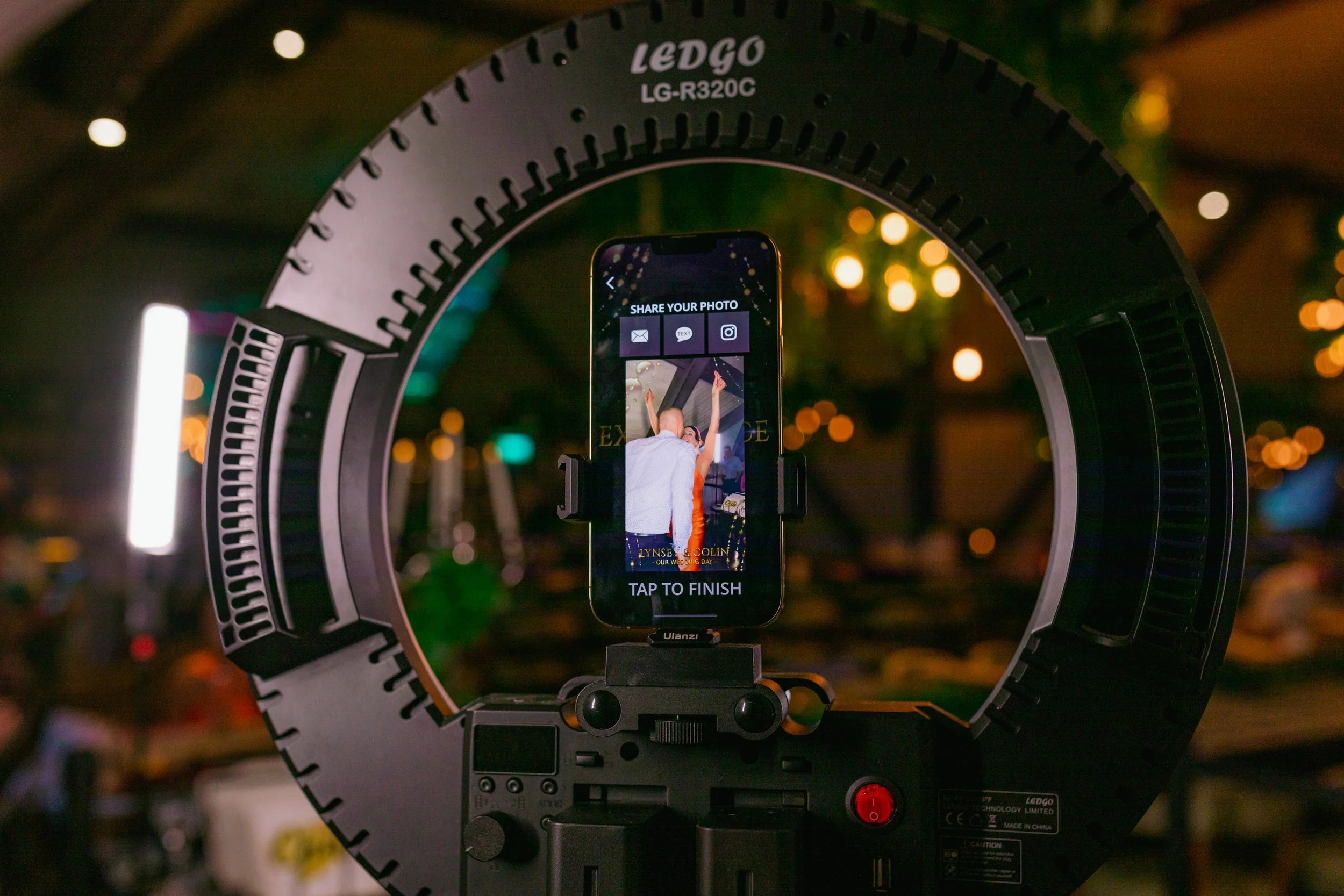 A camera rig with a ring light and a phone capturing a photo of a person at a celebration or event, with blurry colorful lights in the background.