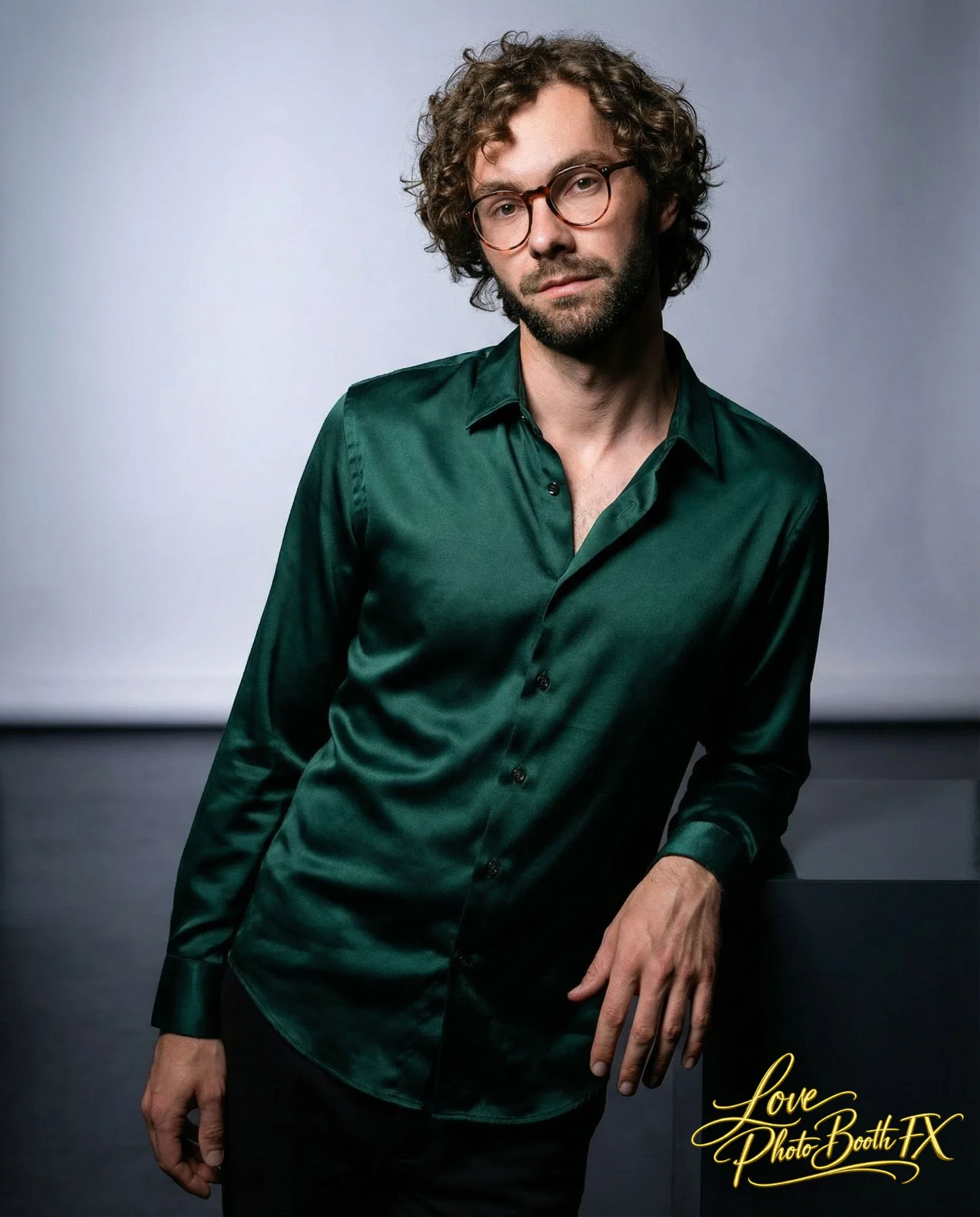 A man with curly hair, glasses, and a beard wears a shiny dark green dress shirt, standing against a plain background.