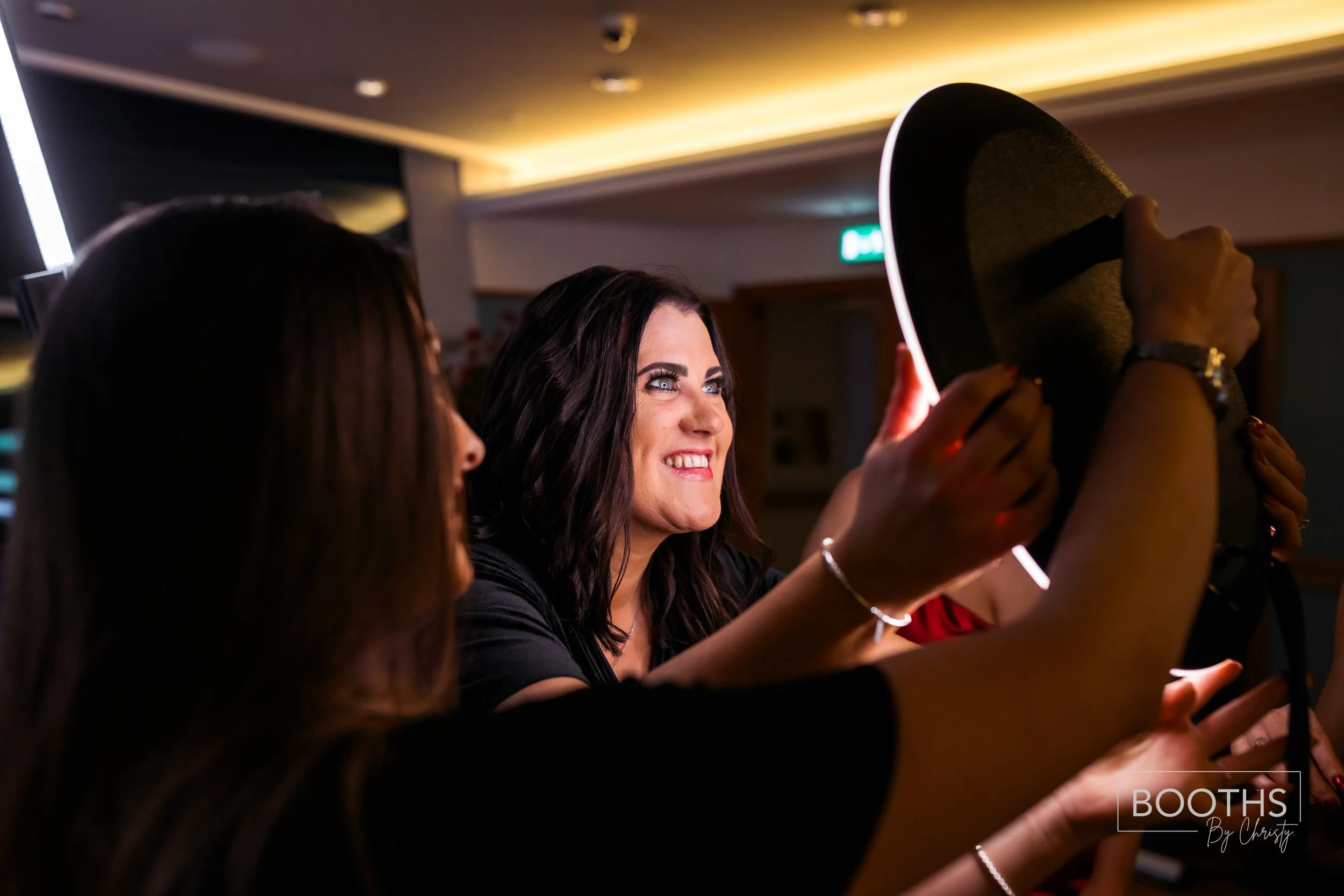 A woman in a black shirt smiles while holding a black hat, fitting it on her head, surrounded by other women in an indoor setting with warm lighting.