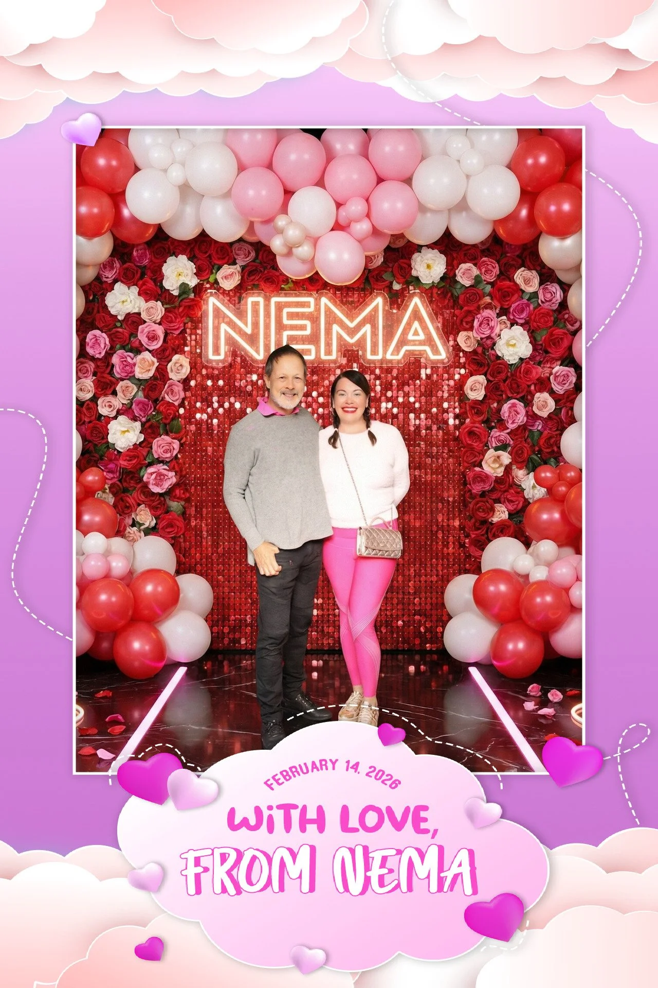 Two people, a man and a woman, standing in front of a decorative backdrop with flowers, balloons, and a neon sign that says 'NEMA'. The woman is wearing pink leggings and a white top, and the man is in a gray sweater. The event date, February 14, 202