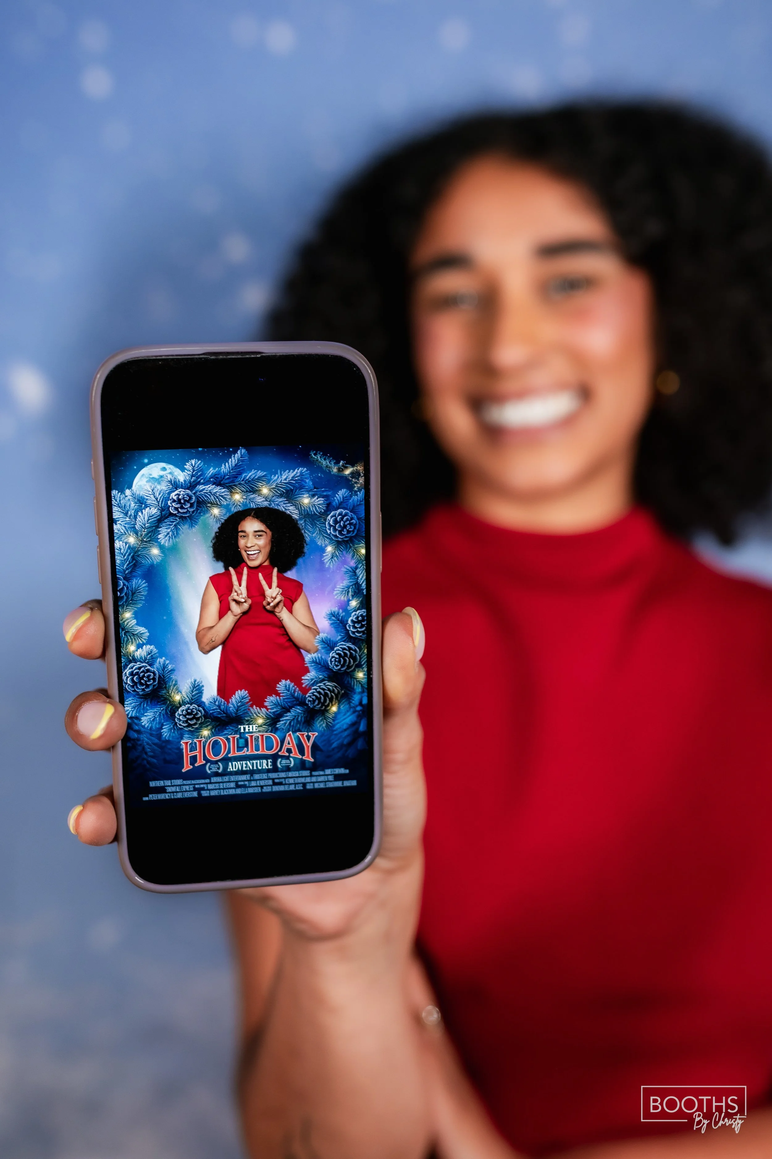 Woman with curly black hair smiling and holding a phone displaying a holiday-themed movie poster, with a festive blue and snowflake background.