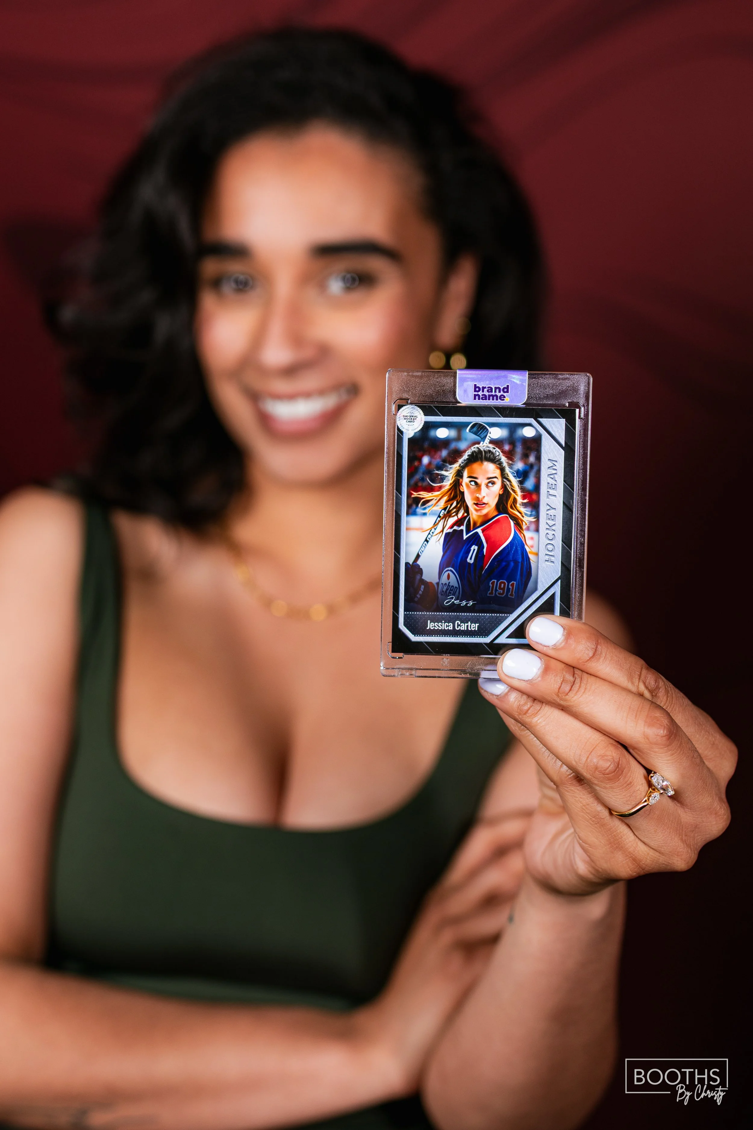 A woman with dark, curly hair smiling while holding a hockey trading card featuring a female hockey player named Jessica Carter.
