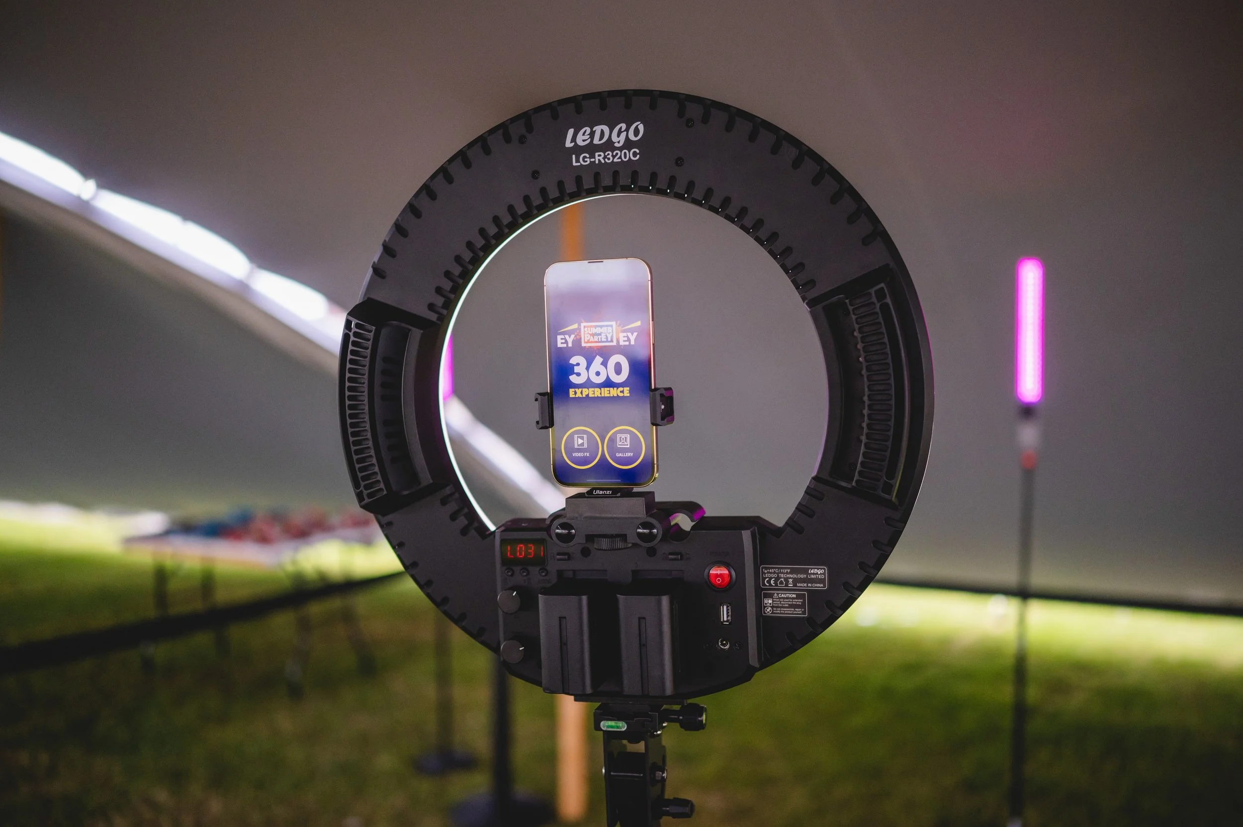 Ring light setup with a smartphone displaying an app for 360-degree video recording, with a blurred background of a bridge and grassy area.