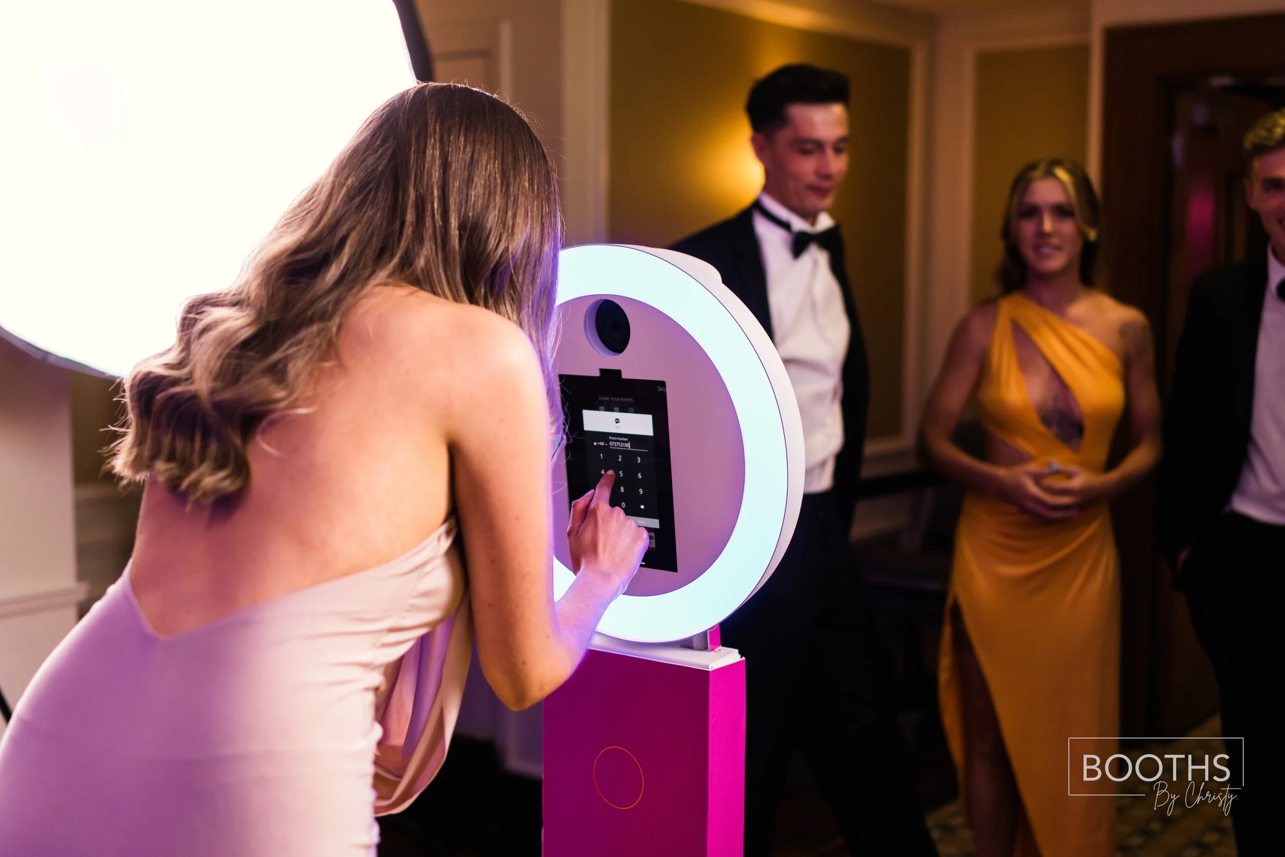 Women using a photo booth at a formal event, with three men and women in formal attire standing in the background.