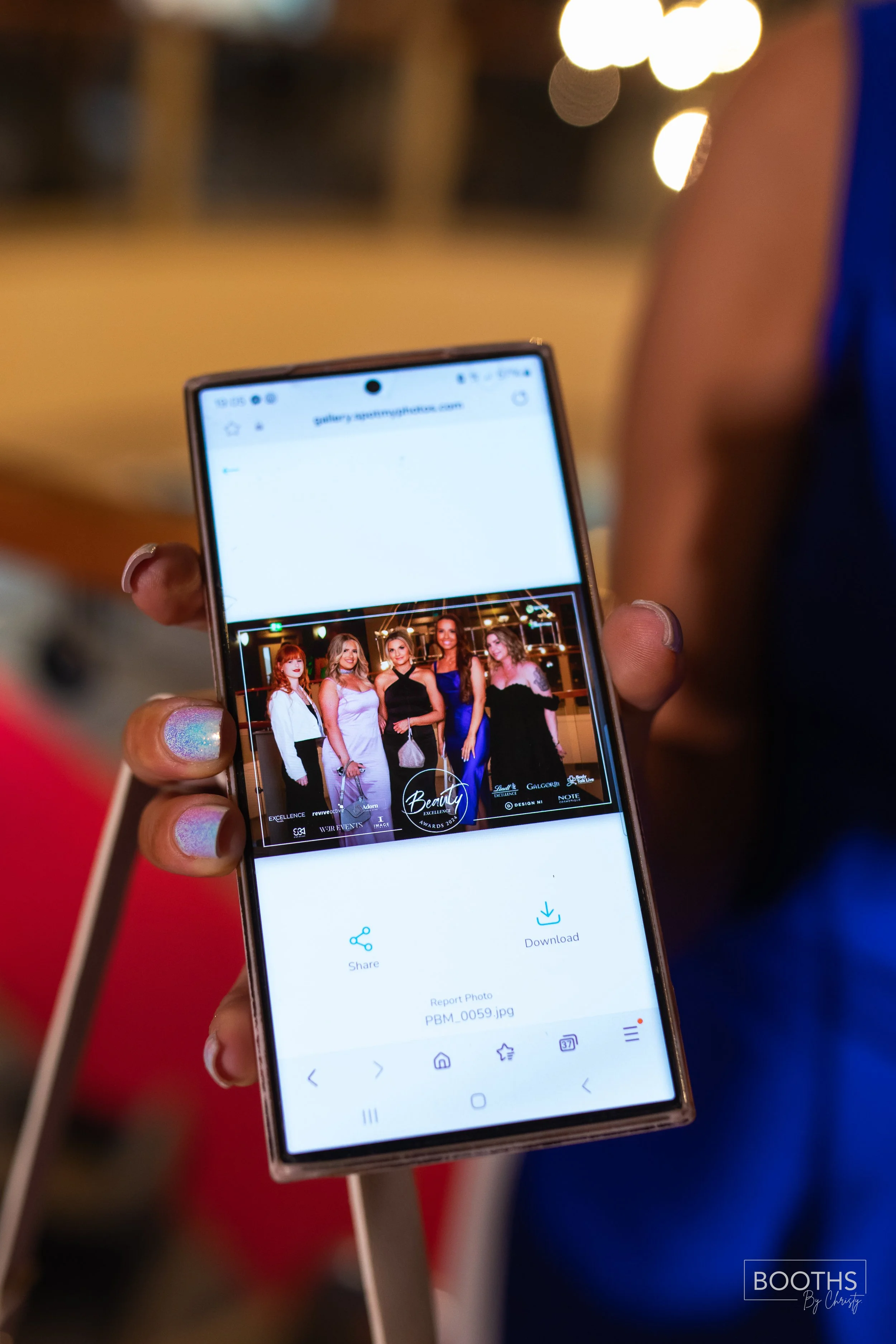 A person holding a smartphone displaying a photo of five women dressed in formal attire at an event, with blurred background lights.
