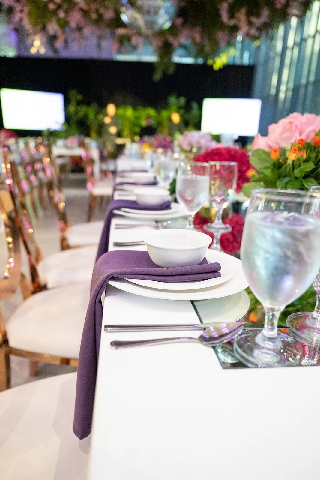 Elegant table setting with white plates, purple napkins, water glasses, and floral centerpieces, arranged in a decorated event space with greenery and monitors in the background.