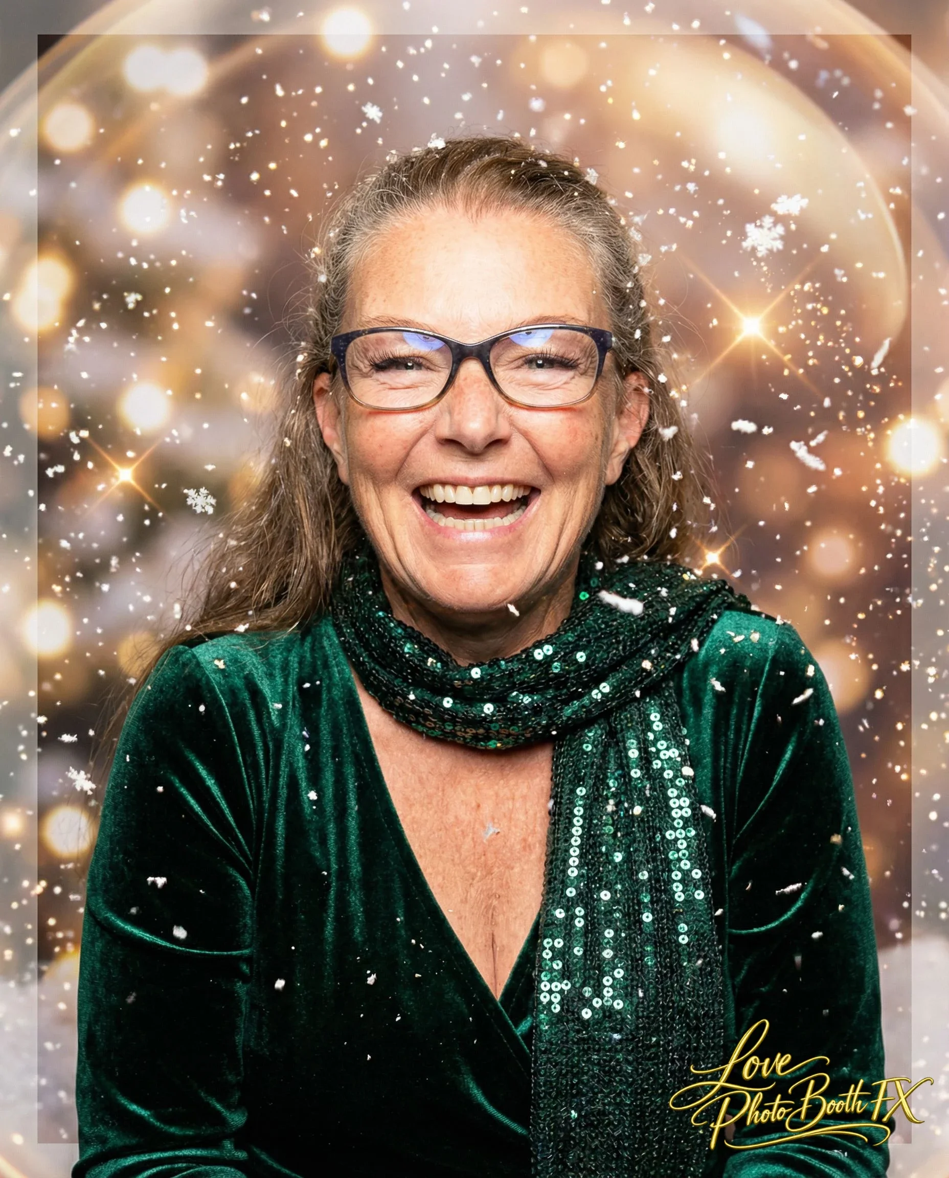 A smiling woman wearing glasses and a green velvet top, with a glittery scarf around her neck, surrounded by a festive sparkly background with snowflakes and glowing lights.