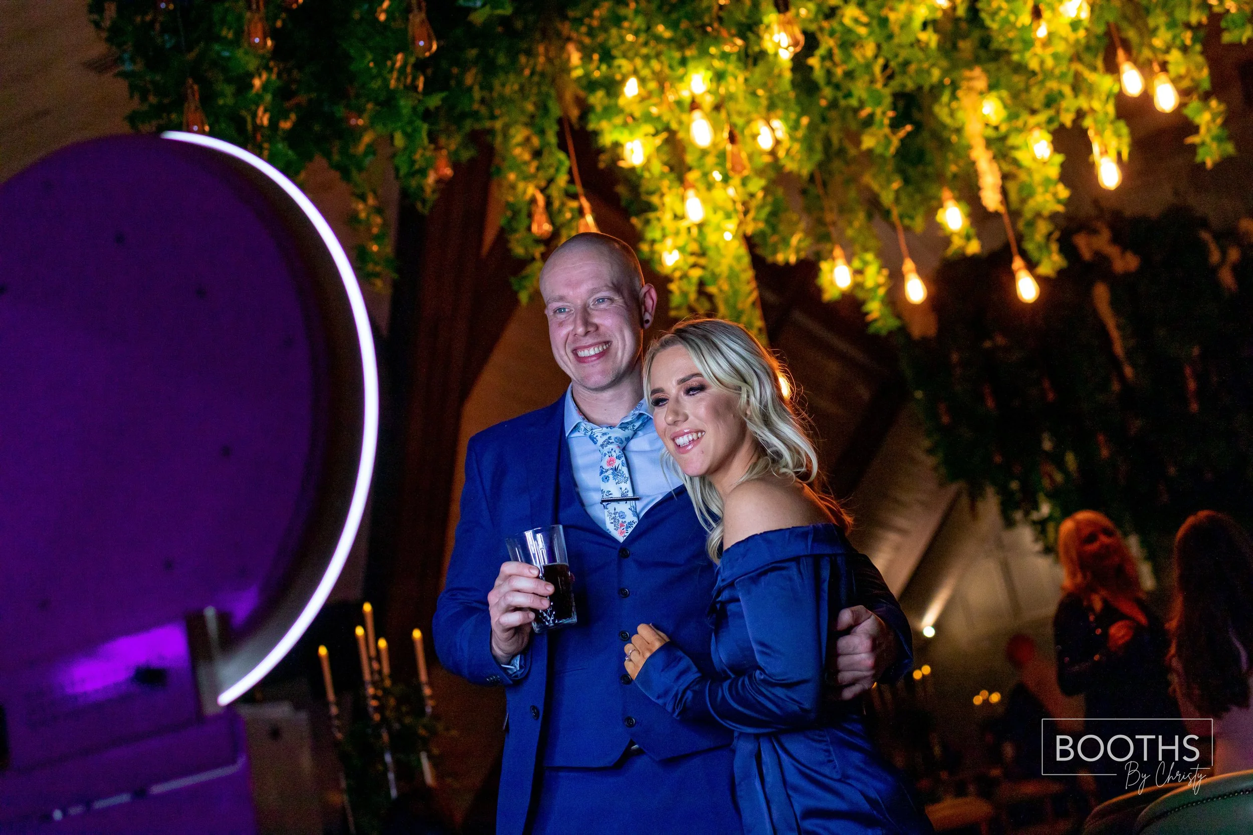 A man and woman smiling and posing at an indoor event, with the man holding a drink, under hanging lights and green leaves.