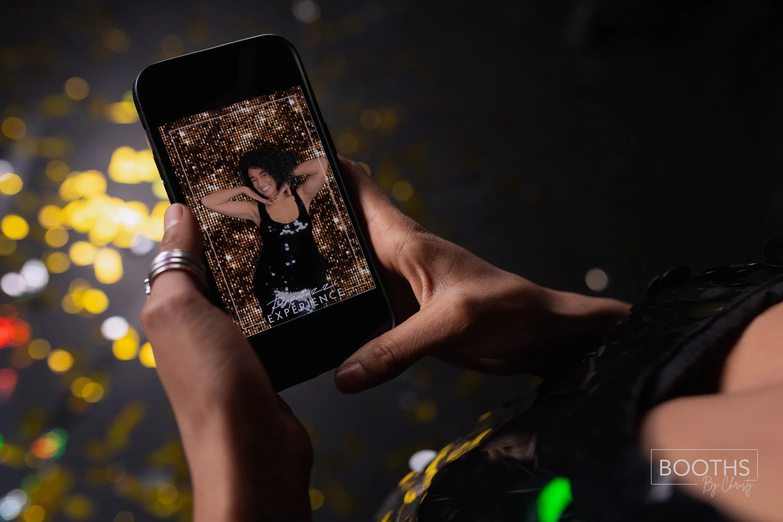 Person holding a smartphone displaying a photo of a woman smiling with arms behind her head, standing against a sparkling gold backdrop.