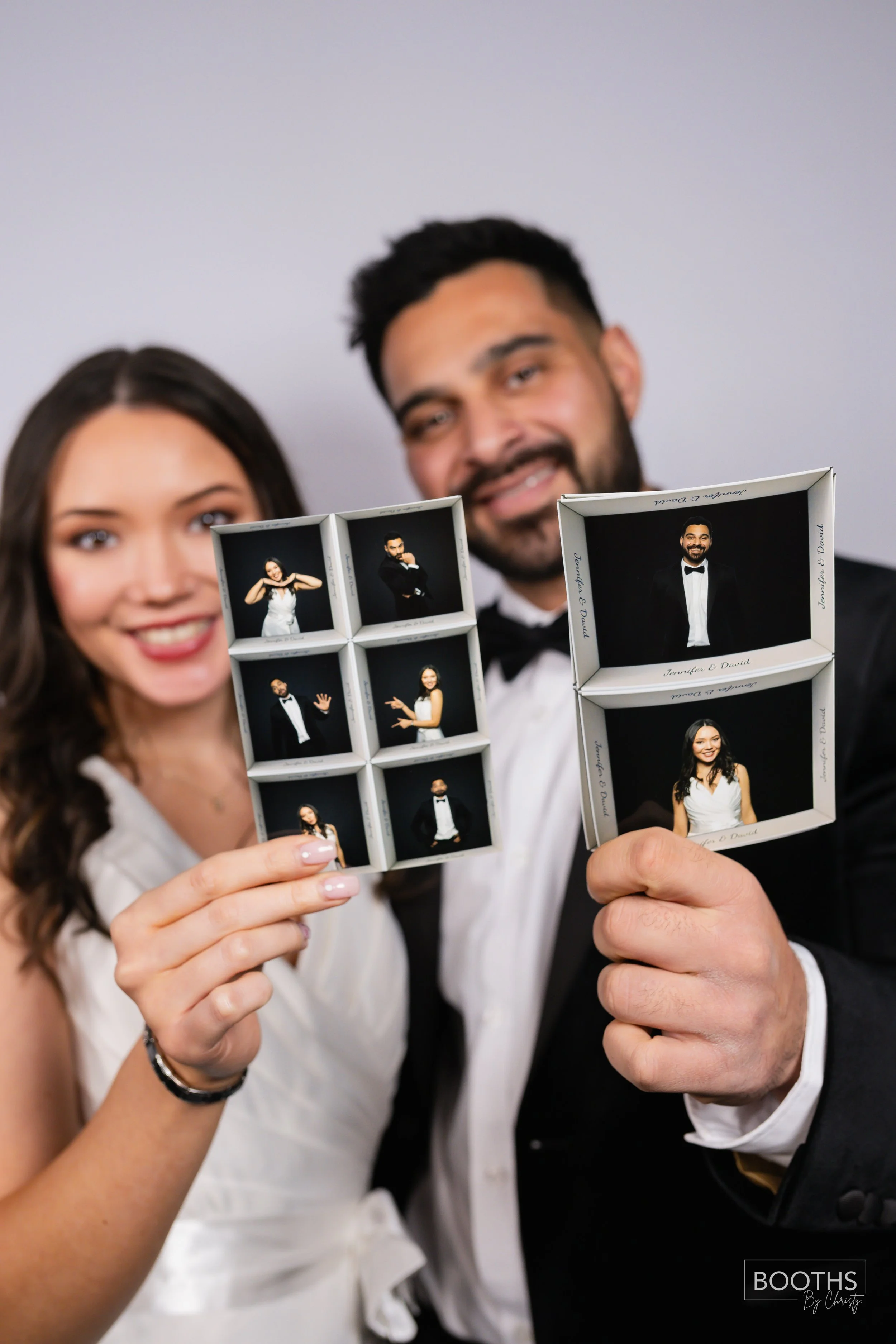 A man and woman in formal attire holding photo booth picture strips at a celebration, smiling at the camera.