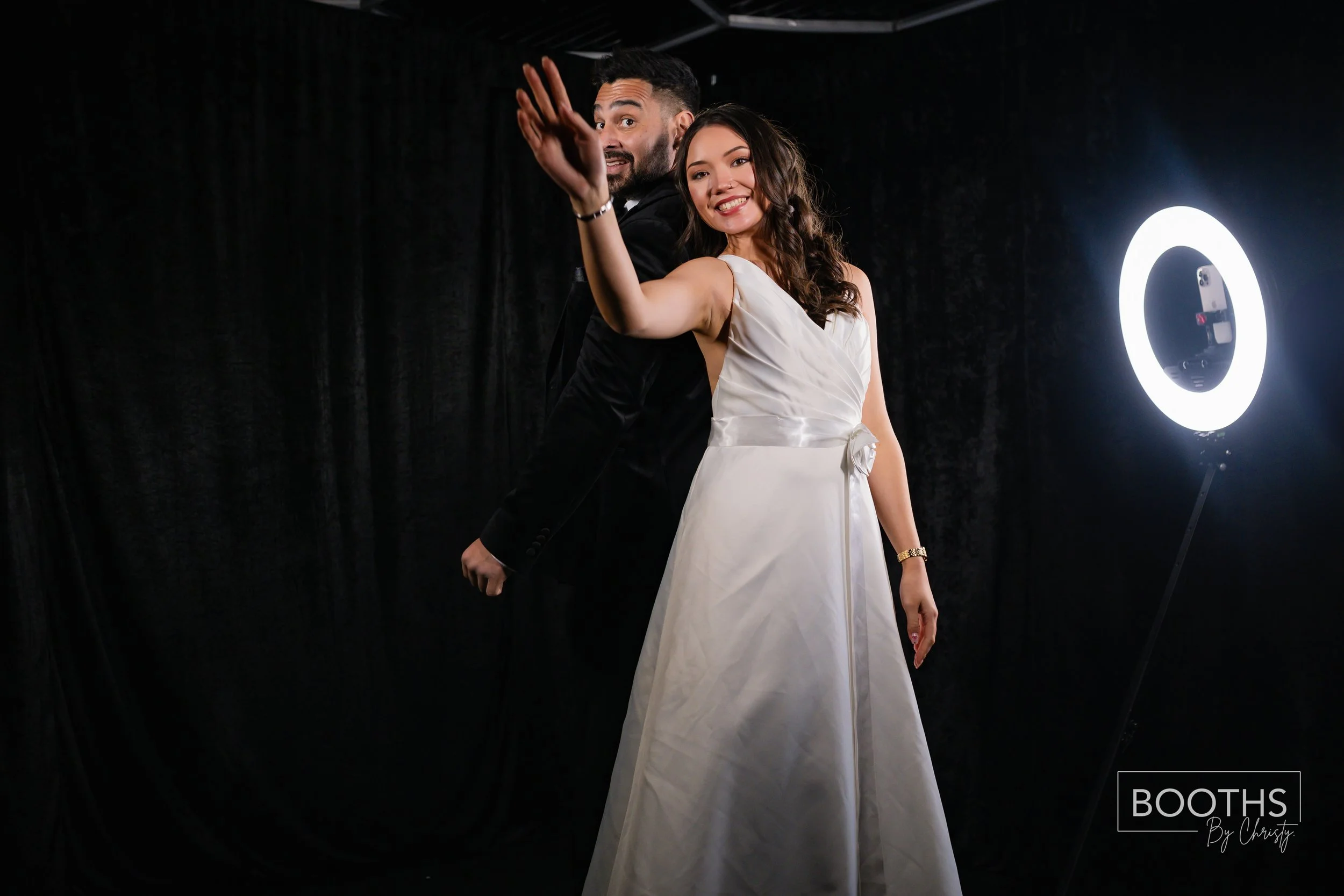 A smiling woman in a white dress and a man in a black suit posing for a photo in a photo booth, with a ring light on the right side of the image.