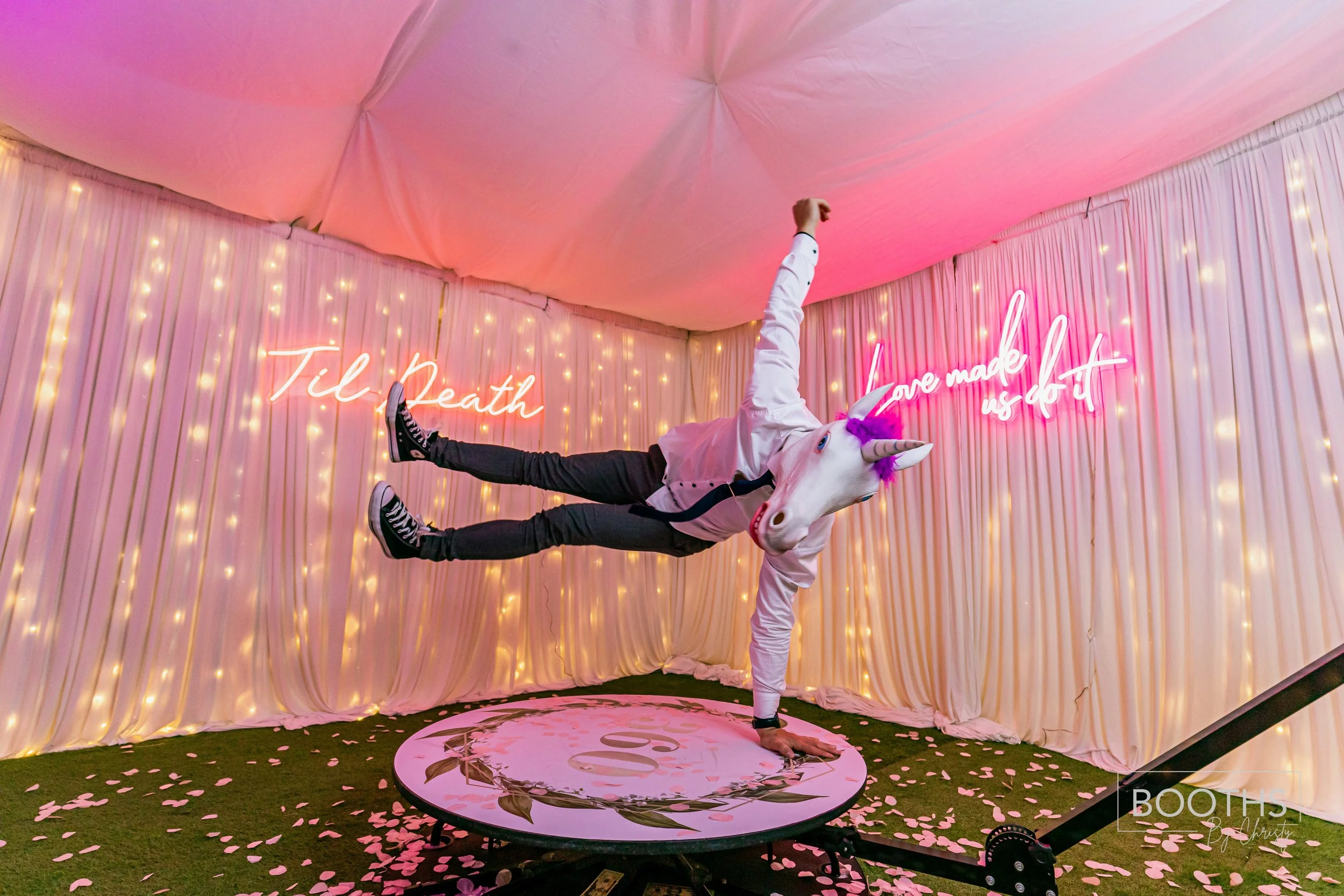 Person wearing a unicorn mask and a white jacket is holding a handstand on a circular platform with pink flower petals scattered on the floor. The background has pink neon signs that say 'Til death' and 'Love made us do it' surrounded by string light