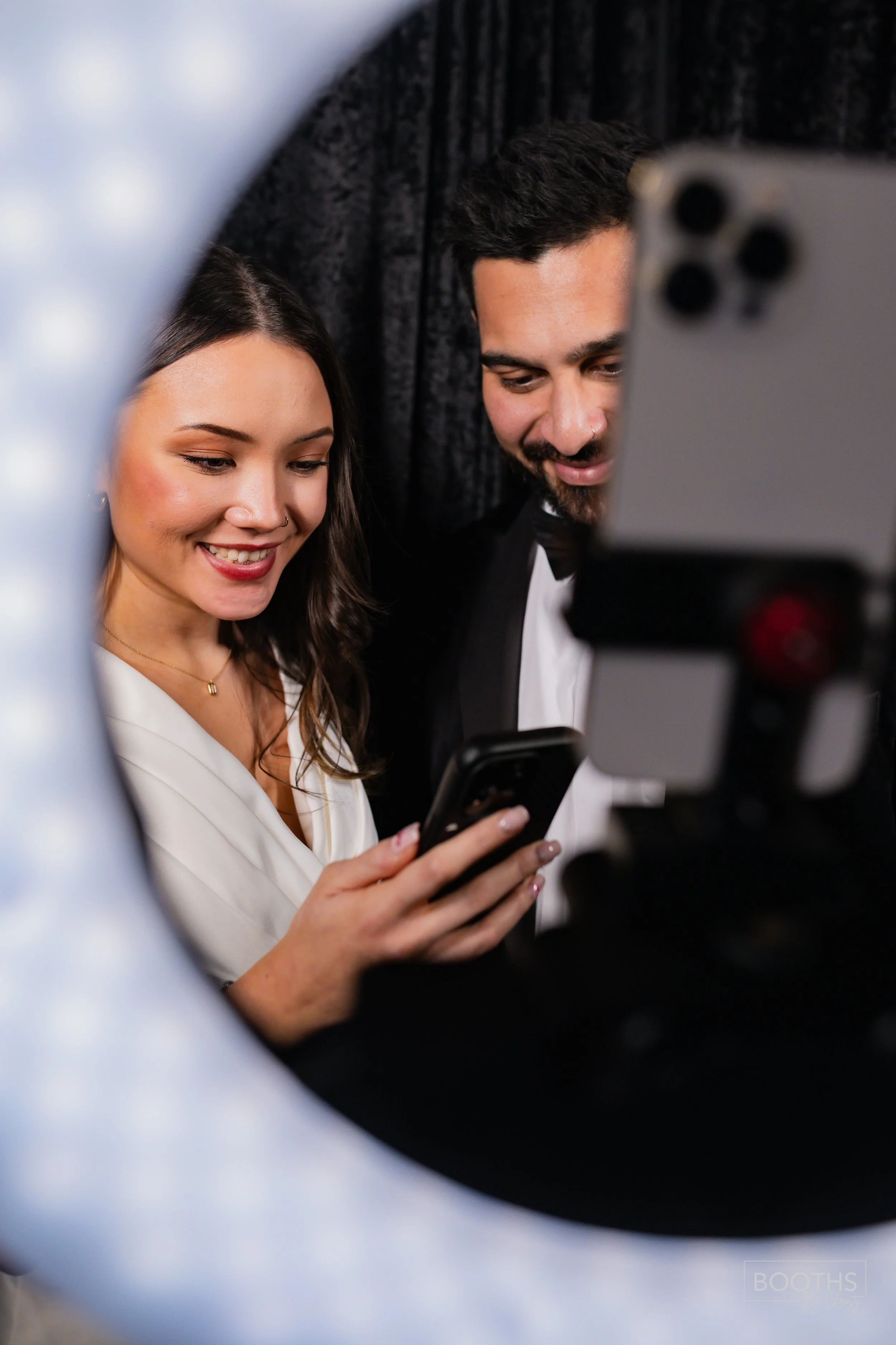 A woman and man dressed in formal attire, smiling and looking at a smartphone, seen through a ring light used for photography.