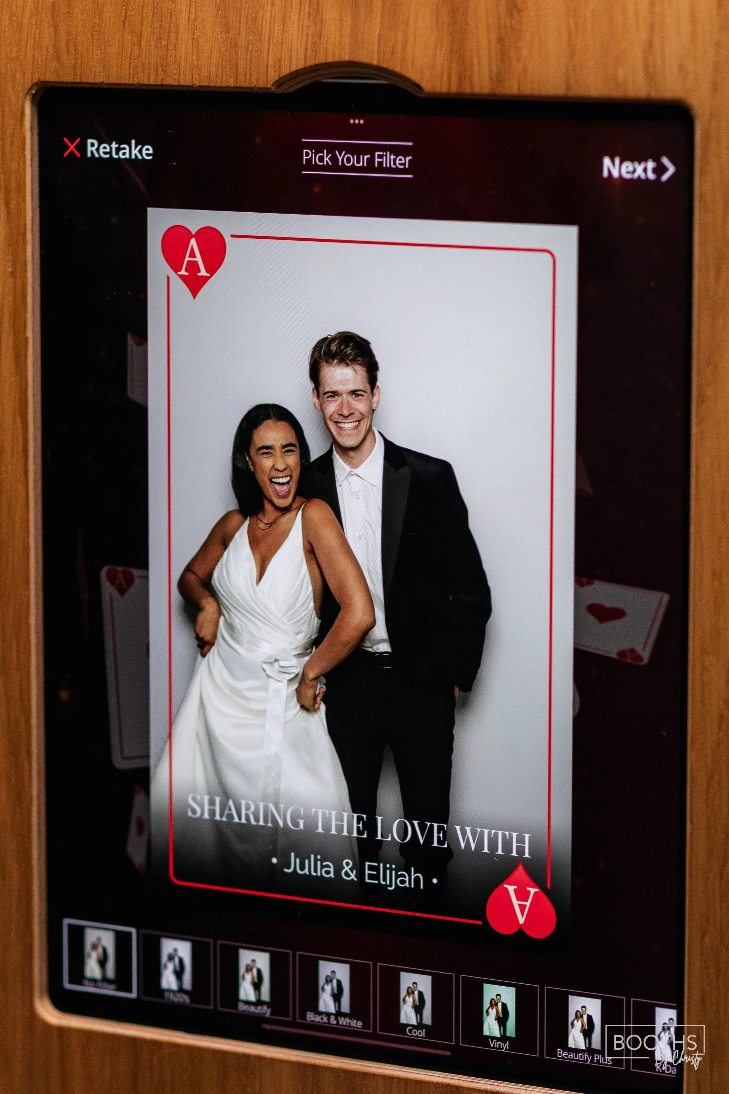 A digital screen displays a photo booth template of a smiling couple, Julia and Elijah, in formal attire, with the text 'Sharing the Love with'. The template has a card suit design with hearts, and the screen shows options for photo filters.