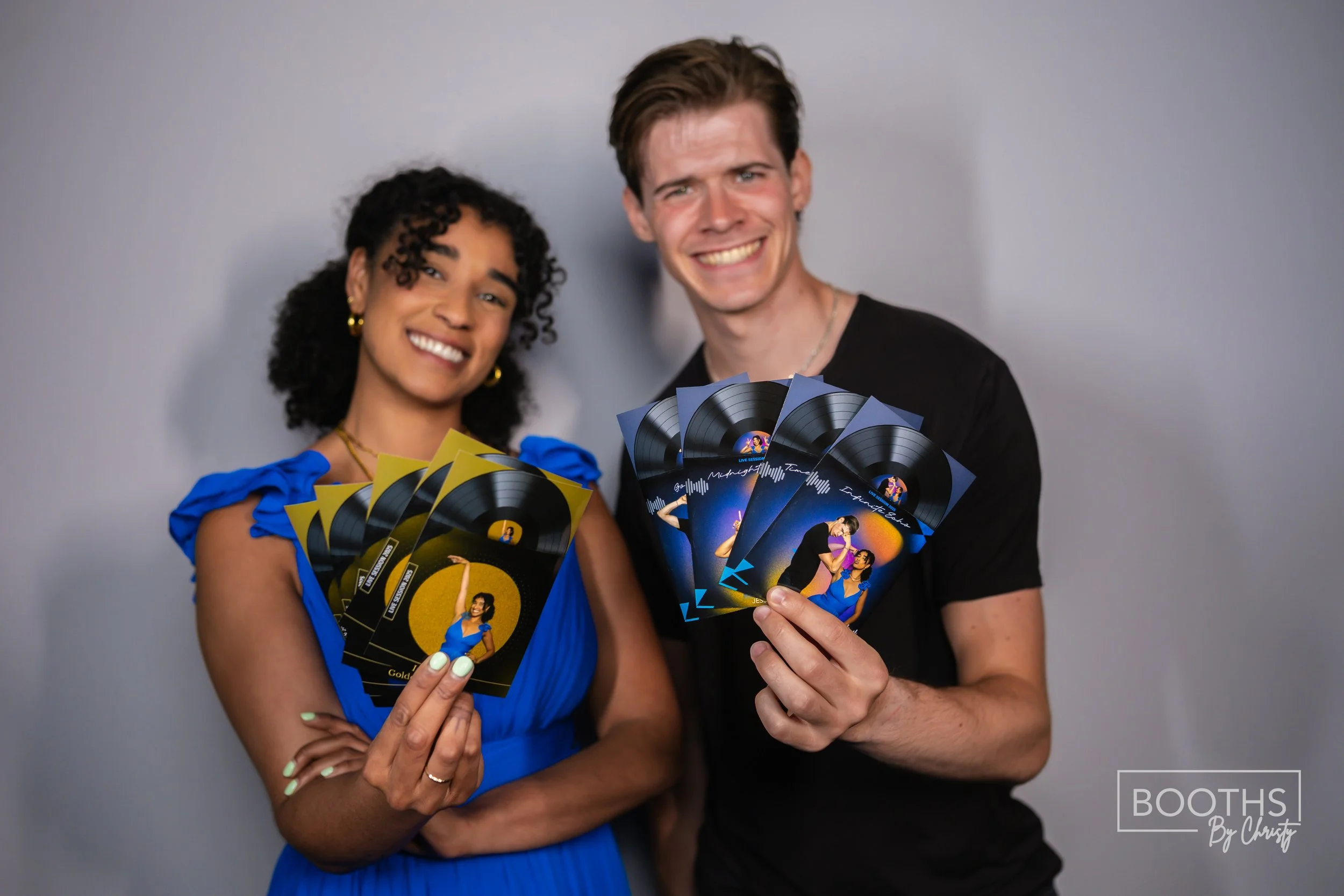A smiling woman in a blue dress and a smiling man in a black shirt holding multiple cards with pictures and graphics, standing against a plain background