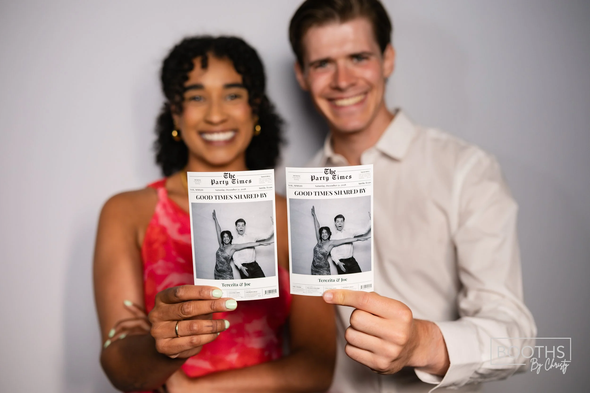 A smiling couple holding up black and white photo booth pictures of themselves, with the woman in a red dress and the man in a white shirt, against a gray background.