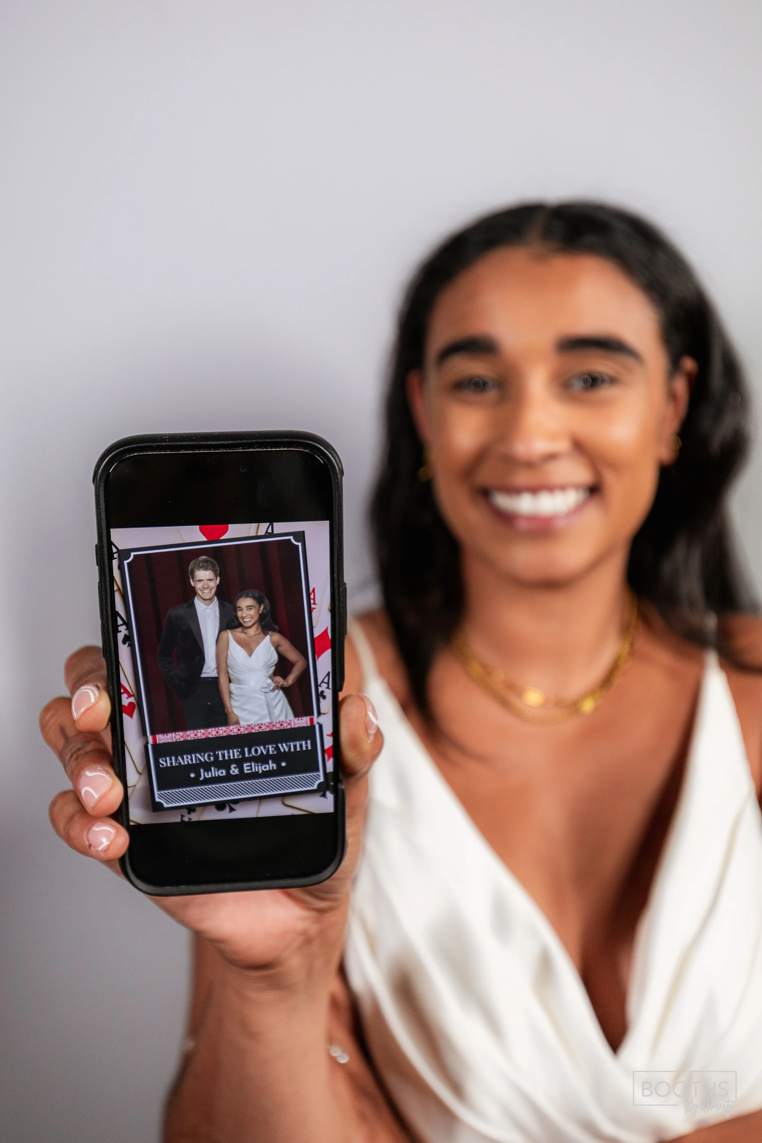 A woman smiling and holding a smartphone displaying a photo of a couple dressed in formal attire with a caption reading 'Sharing the Love with Julia & Elijah'.