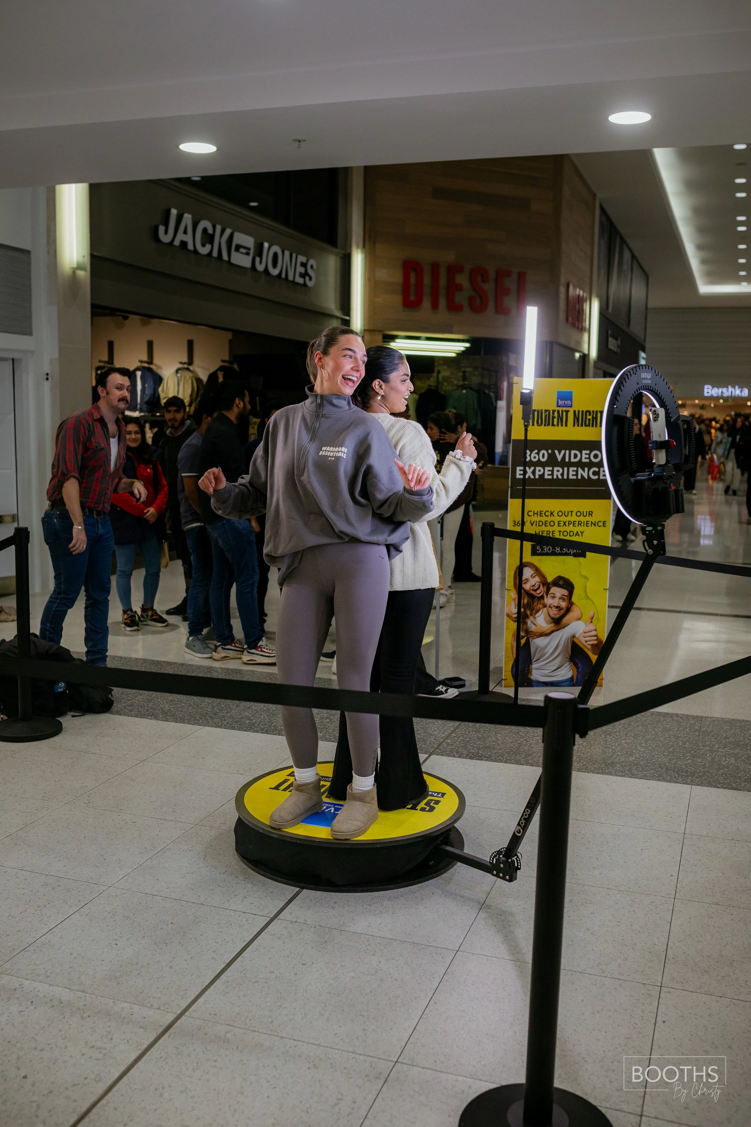 Two women are standing on a rotating platform at a shopping mall, taking a photo with a 360-degree camera. One woman is wearing a gray sweatshirt and matching sweatpants, while the other is in a cream-colored sweater and black pants. There is a line