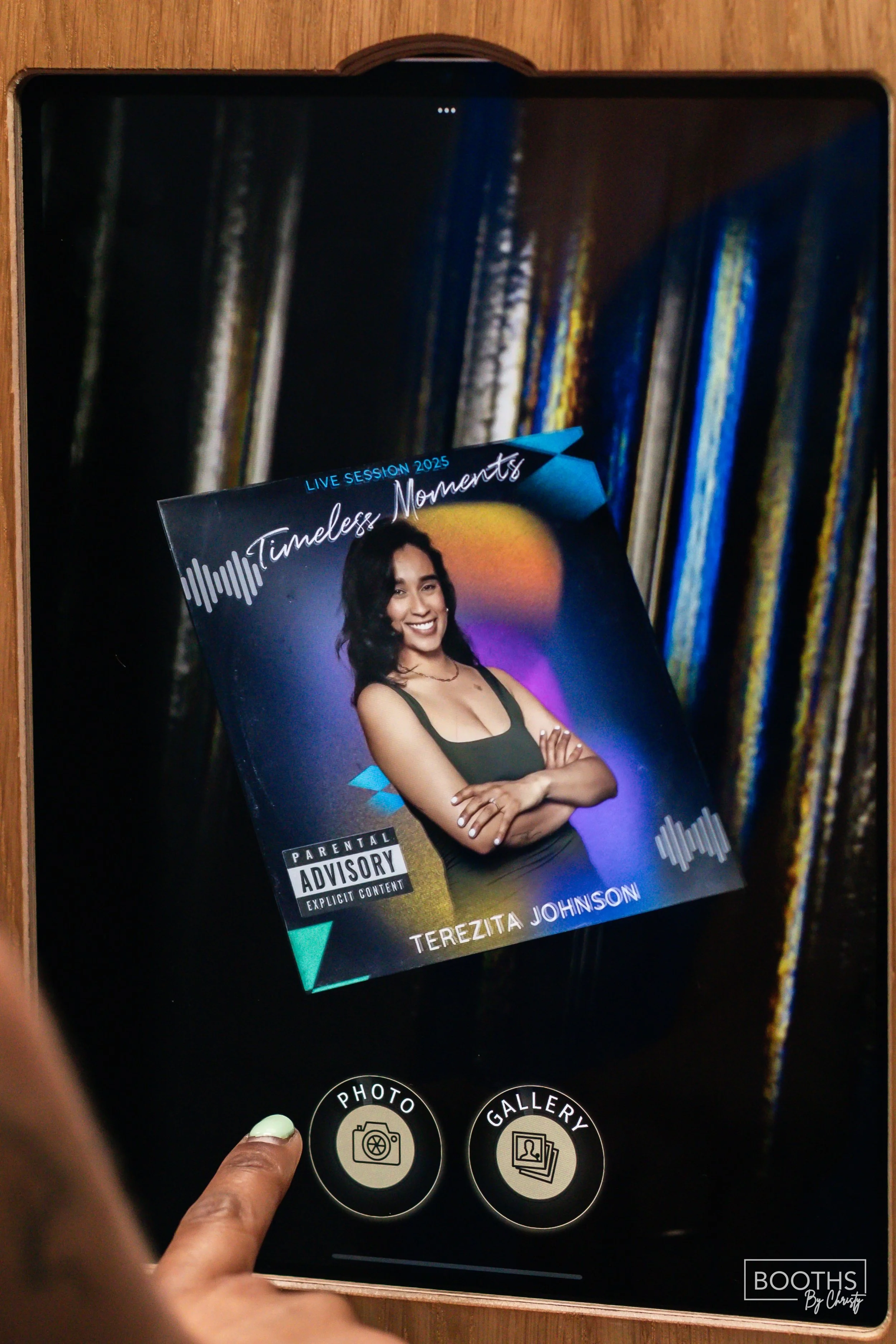 A person's hand is holding a tablet displaying a photo of an album cover titled "Timeless Moments" by Terezita Johnson, featuring a woman smiling with crossed arms against a sunset background, with options for taking a photo or viewing a gallery belo
