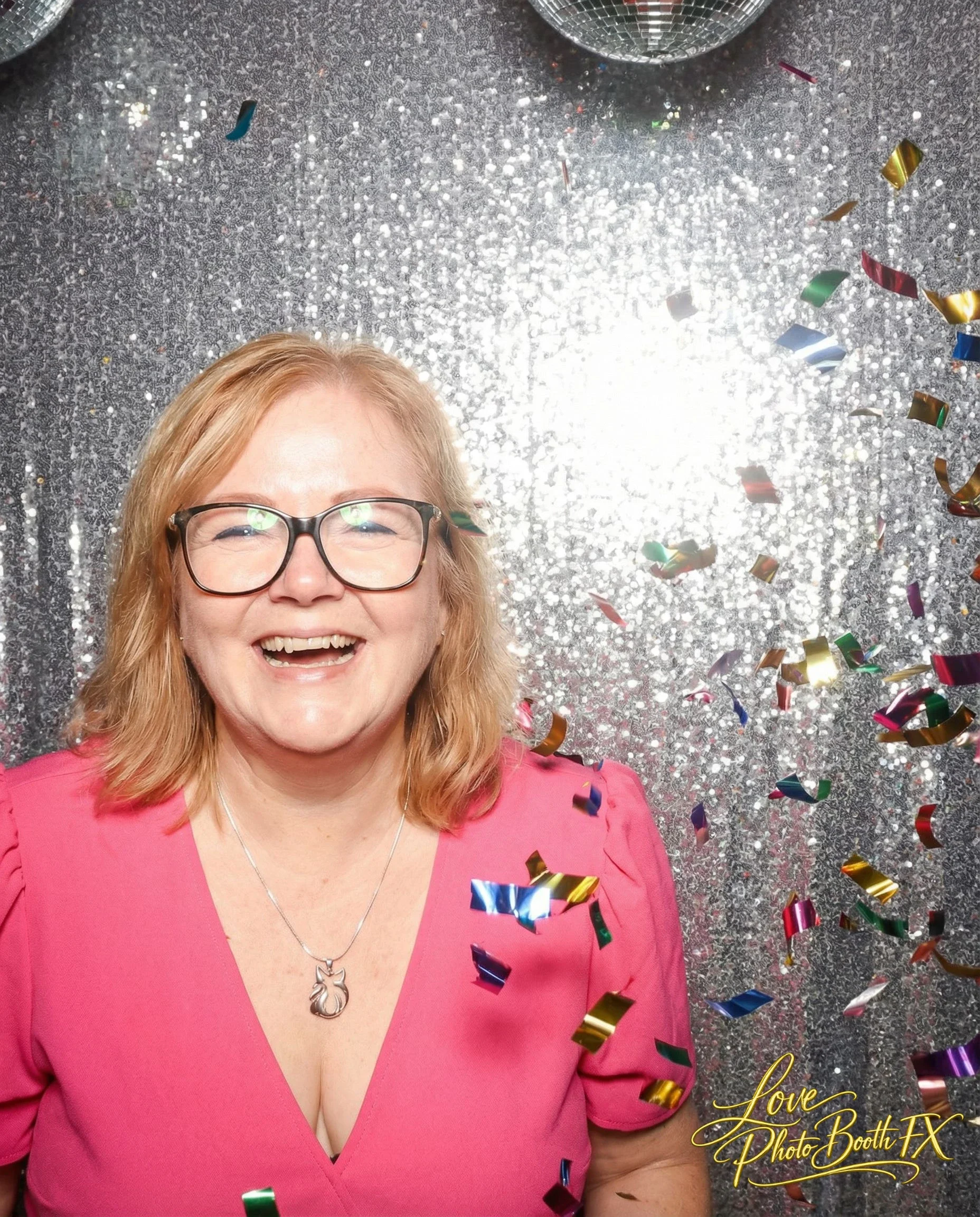 A woman with glasses smiling in front of a silver, glittery backdrop with colorful confetti falling around her.