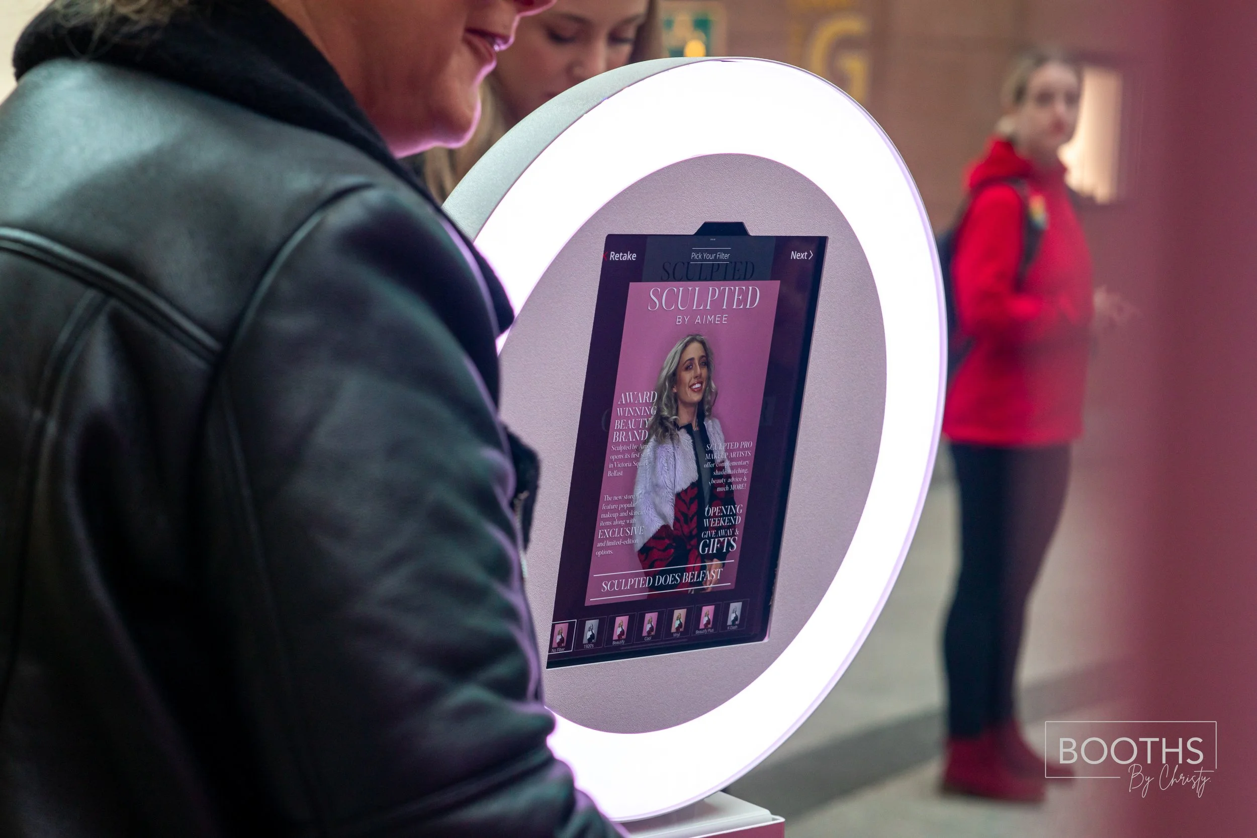 Person using a digital makeup mirror at an event, with another person in the background wearing a red hoodie.