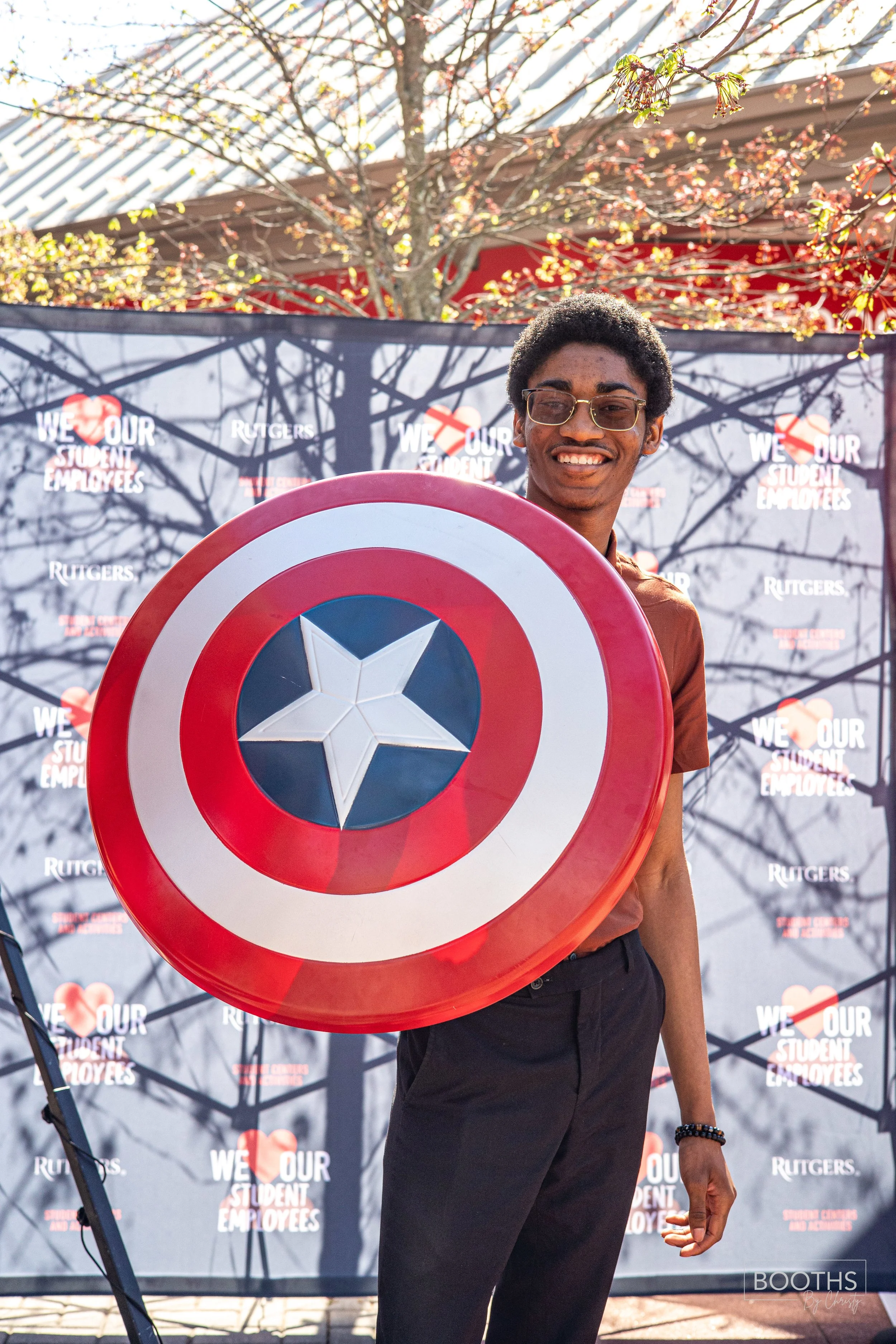 A young man smiling and holding Captain America's shield during a Rutgers event, with a backdrop that says "We Love Our Student Employees" and a tree with budding leaves in the background.