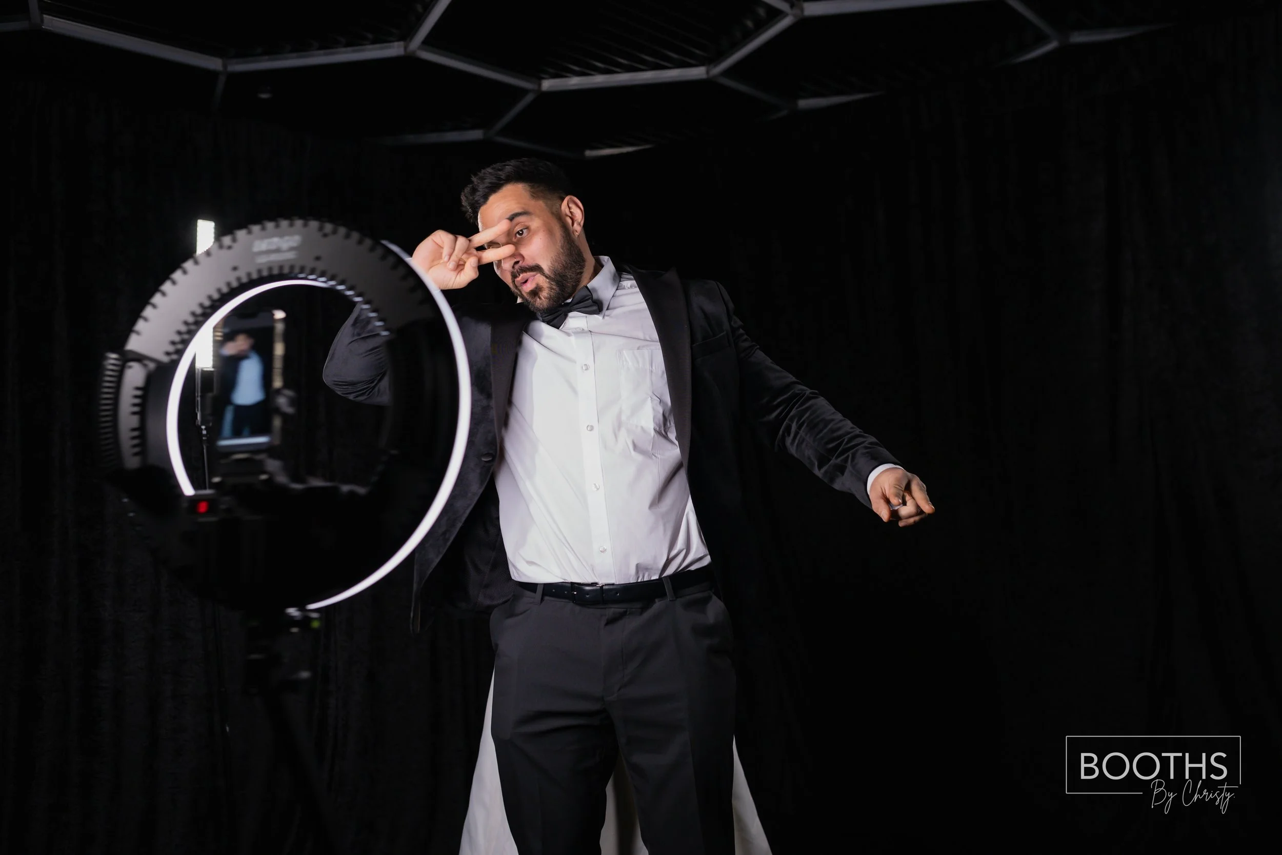 Man in tuxedo posing for a photo at a photo booth, with a professional camera and ring light setup, wearing a white shirt, black bow tie, and black blazer, making a peace sign near his eye.