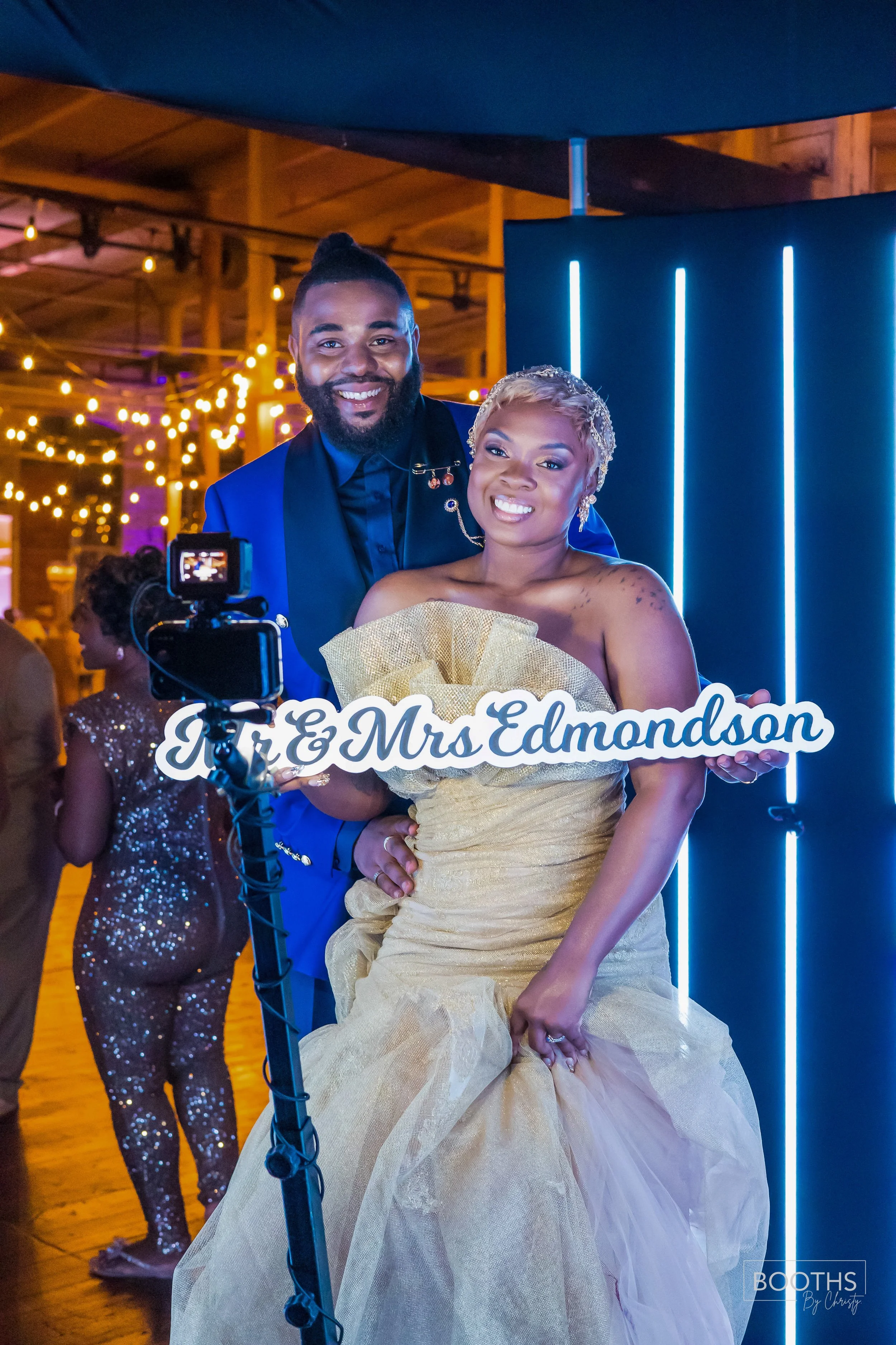 Photo of a smiling couple at a wedding celebration, with the woman holding a sign that reads 'Mr & Mrs Edmondson', in a decorated venue with string lights and blue neon lights in the background.
