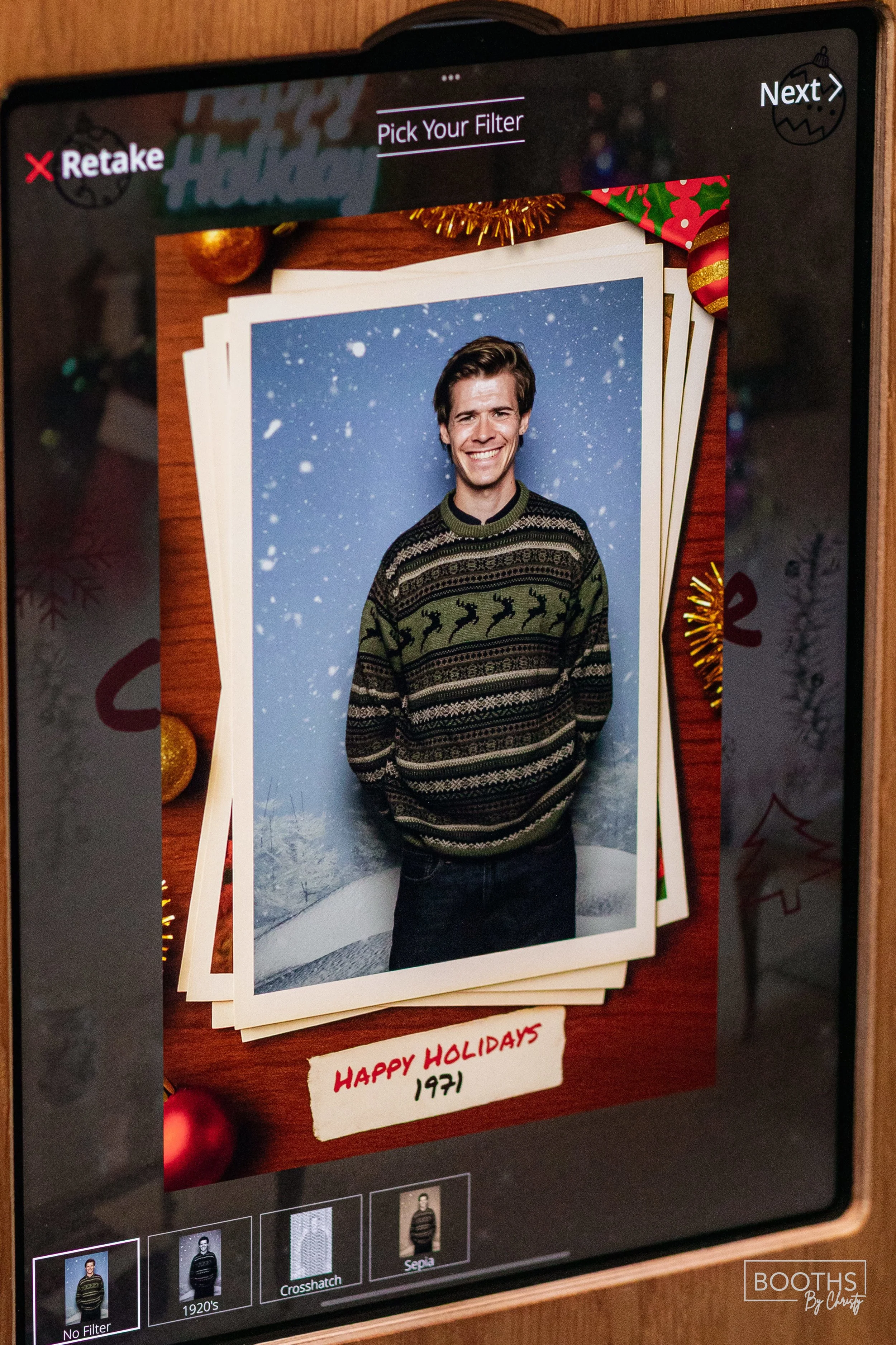 A man smiling in a holiday-themed photo booth with a snowy background, wearing a Christmas sweater with reindeer and snowflake patterns. The display screen shows options for filters, with a handwritten note underneath that reads "Happy Holidays 1971.