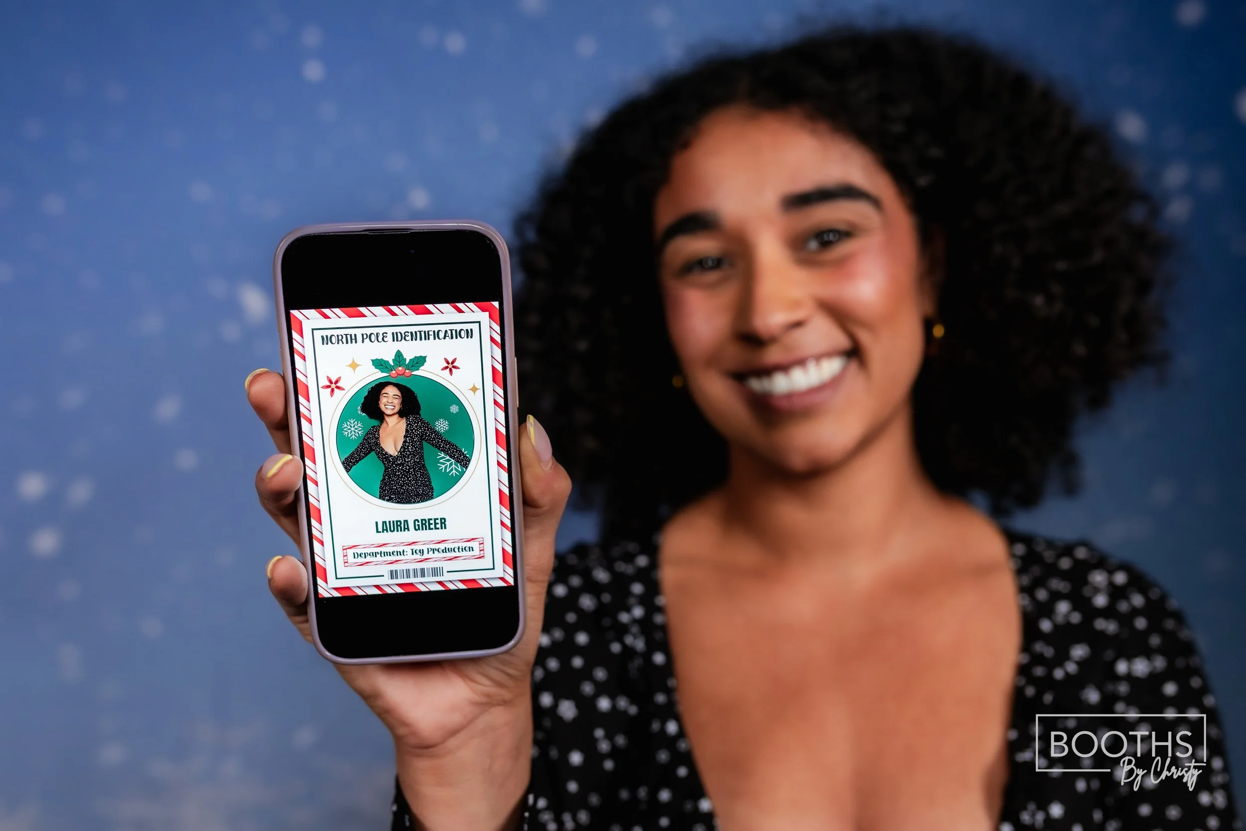A woman smiling and holding a smartphone showing a festive identification badge for Laura Greer with a holiday-themed design, against a blue background with snowflake patterns.