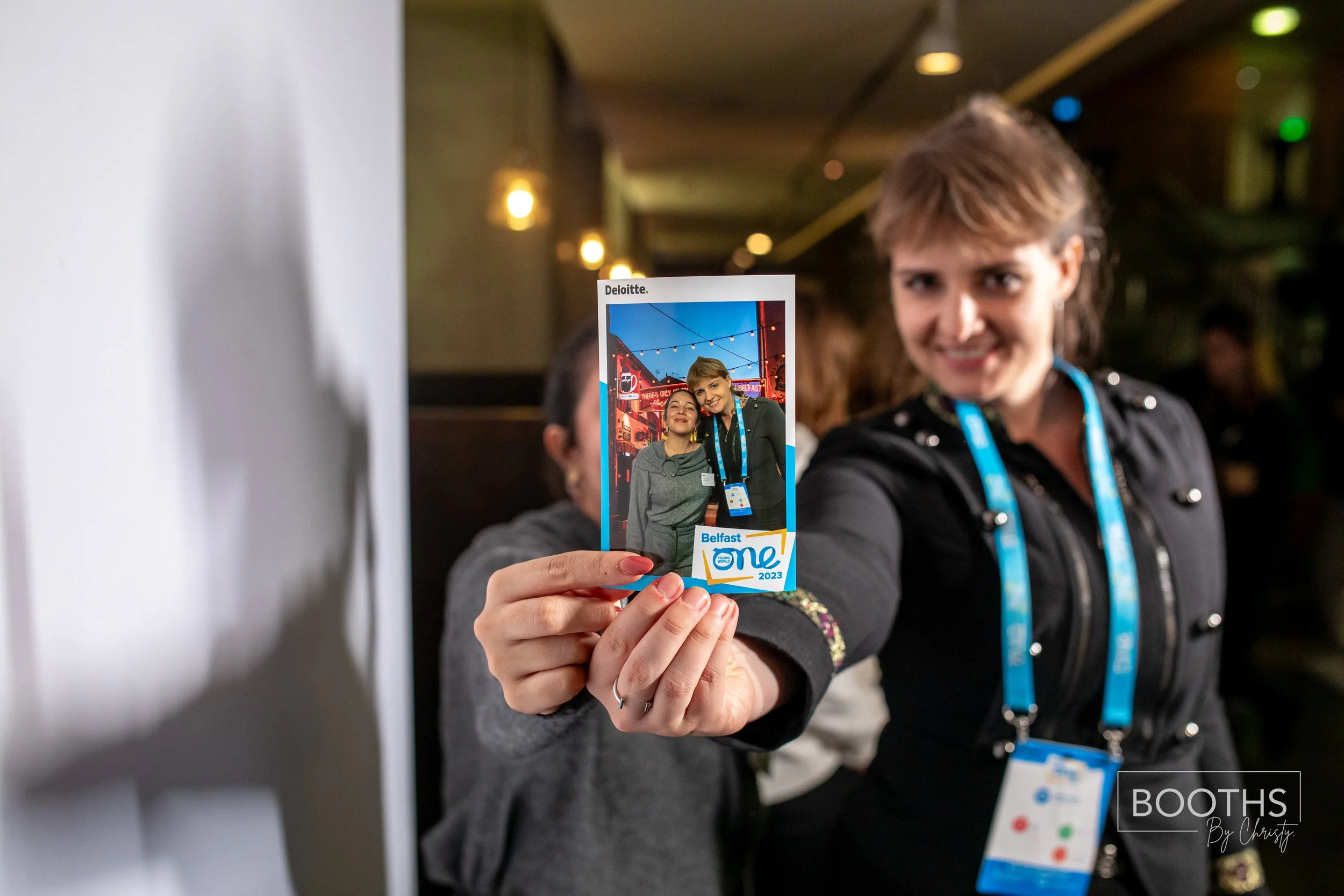 A woman smiling and holding up a badge with a photo of herself and another person at an event called Belfast One 2023.