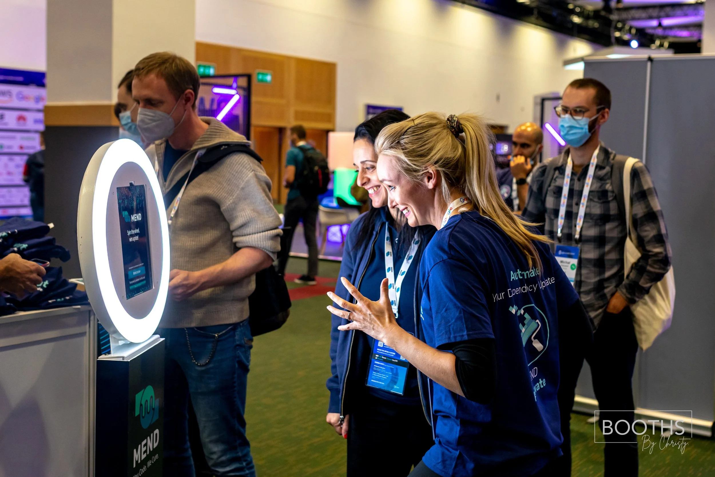 Three women and two men at a conference, engaging with a tech exhibit. One woman is explaining something to others near a circular touchscreen device. All are wearing face masks and conference badges.
