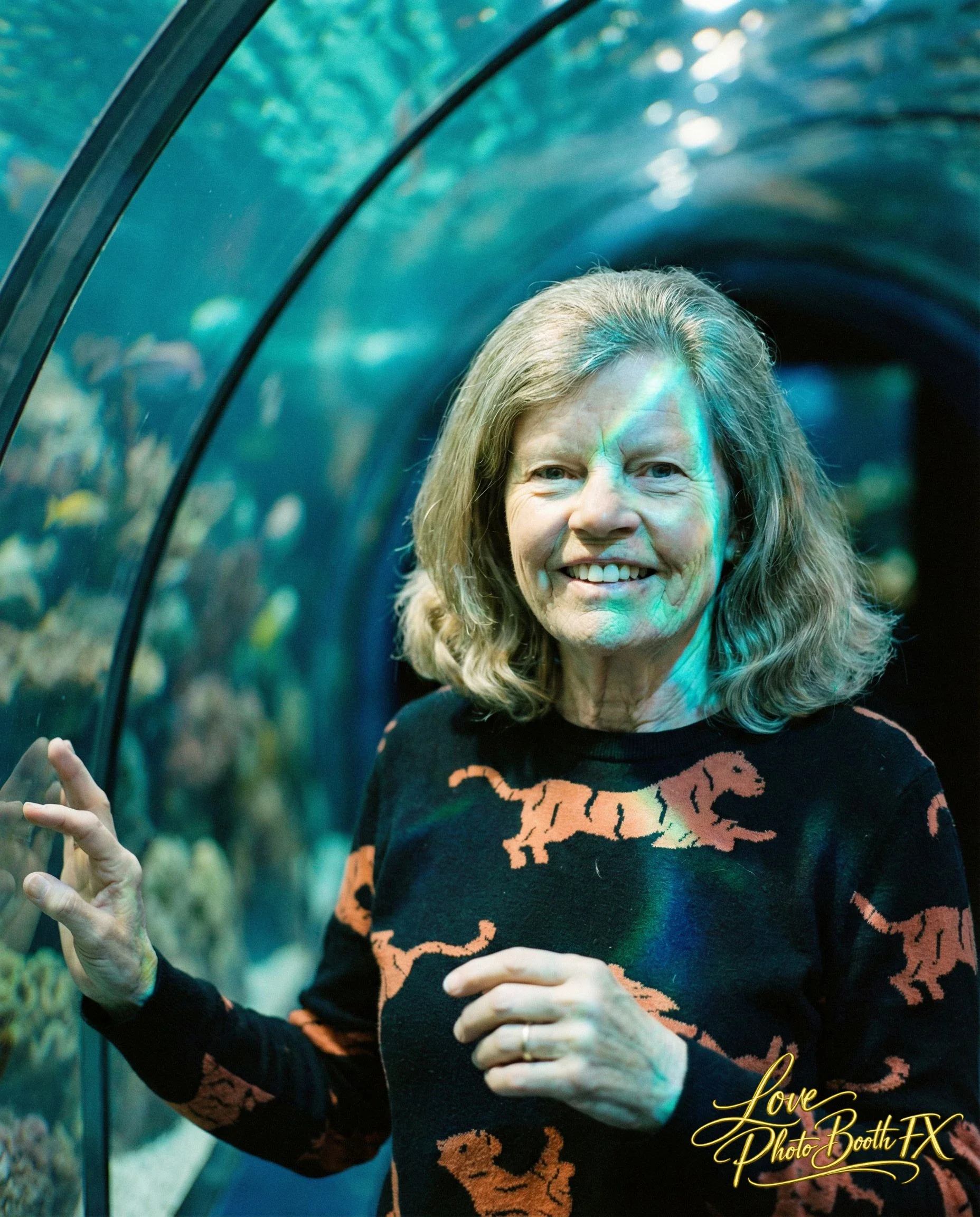 Smiling woman with shoulder-length blonde hair in an underwater tunnel at an aquarium, wearing a black sweater with orange tiger print.