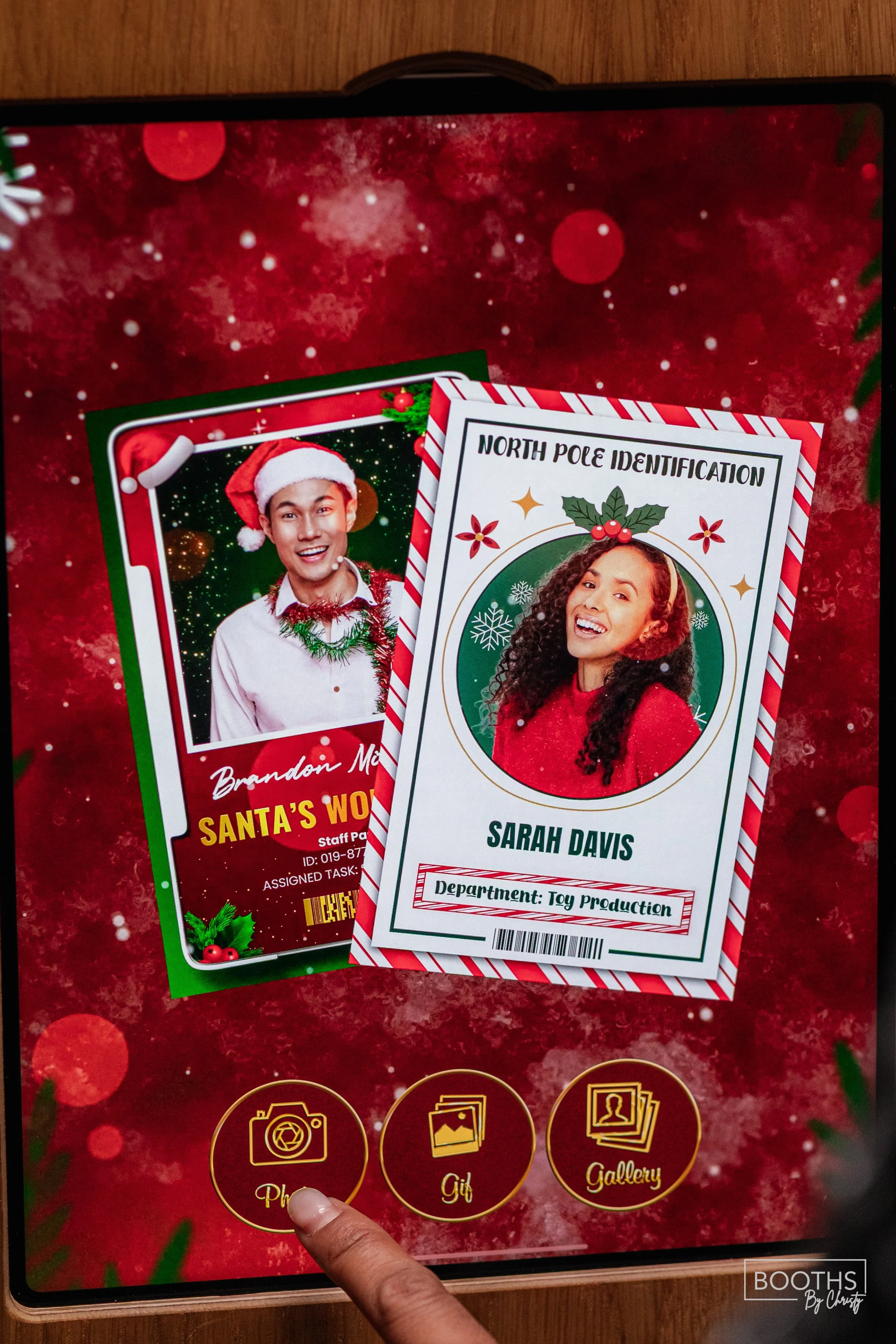 Digital Christmas-themed photo booth screen featuring a Santa's workshop staff card for Brandon M. and a North Pole identification card for Sarah Davis. The screen has icons for photo, gift, and gallery options at the bottom.